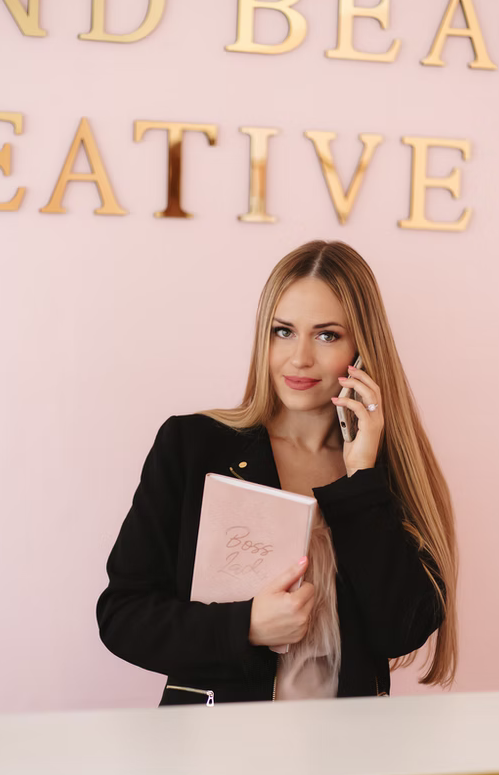 A young woman with long blonde hair, wearing a black jacket, is talking on the phone while holding a pink planner that says 'Boss Babe.' She is standing in front of a light pink wall with large gold letters that spell out words including 'CREATIVE.'