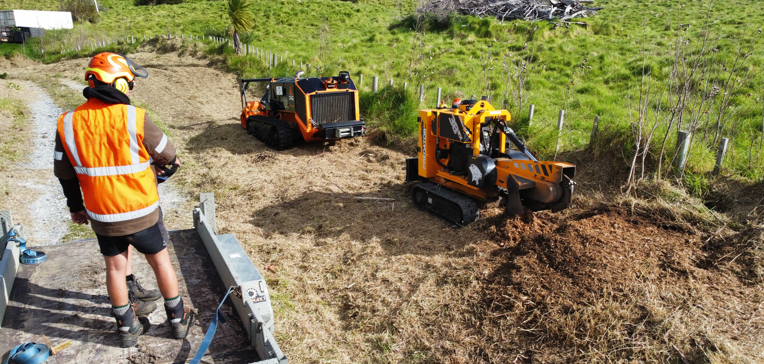 Person in safety vest and helmet operating machinery on a grassy hillside, with a tracked excavator and other construction equipment nearby.
