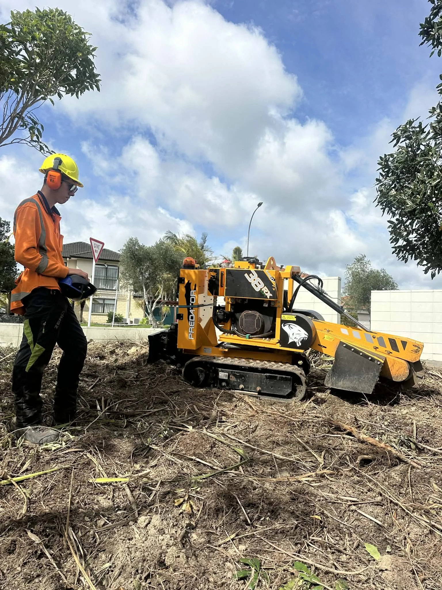 A construction worker wearing an orange safety jacket, yellow helmet, and ear protection operating a yellow tracked stump grinder on a patch of dirt outdoors with trees and residential buildings in the background.