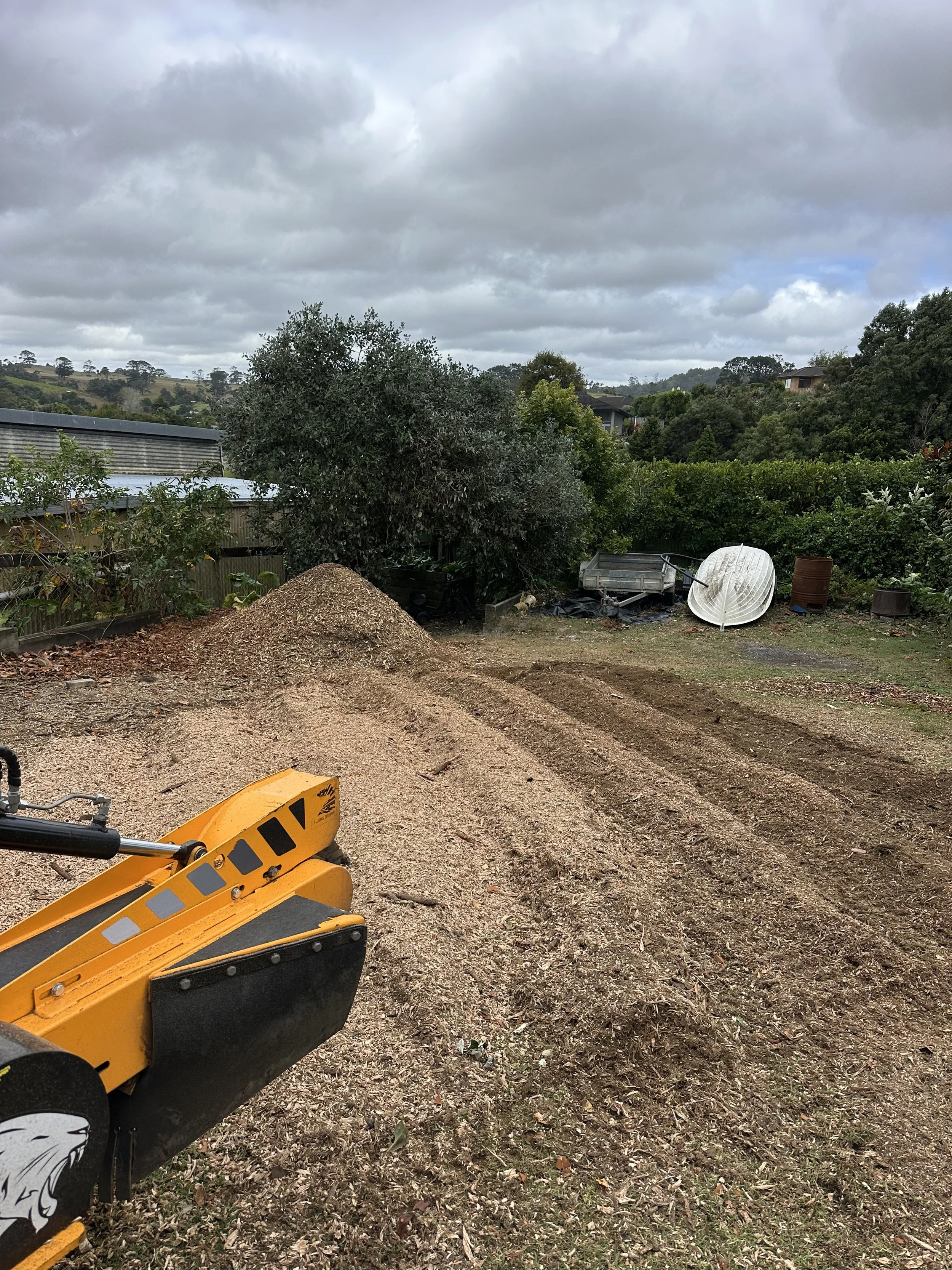 A yard with a yellow stump grinder in the foreground, a pile of mulch, a tree, a boat, a trailer, and various garden items under a cloudy sky.
