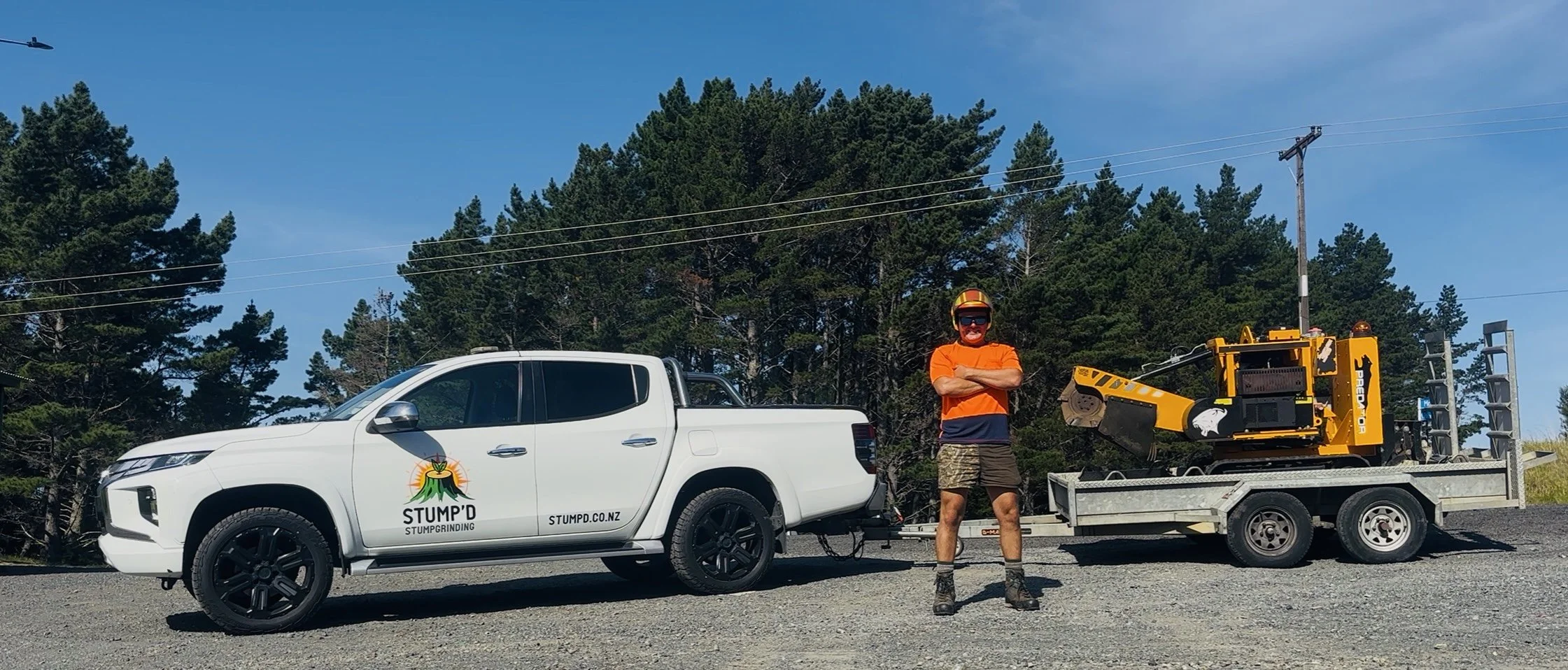 White ute with a logo and the words "STUMP'D" and "stumpd.co.nz" on the door. The truck is on a trailer carrying a remote controlled track grinder.