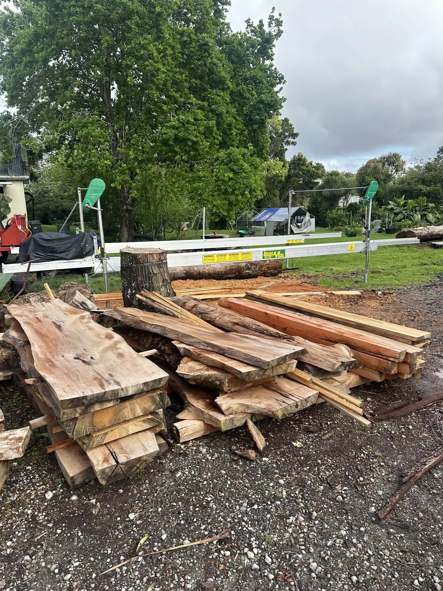 Stacked rough-cut wooden planks and logs outdoors, with a tree stump in the center, in a yard with trees and cloudy sky.