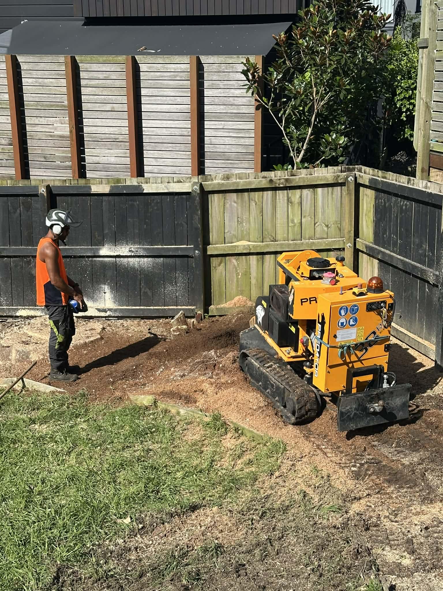 Construction worker wearing safety goggles and helmet stands near a small yellow tracked stump grinder machine in a backyard with a dirt patch, surrounded by wooden fences and a grassy area.