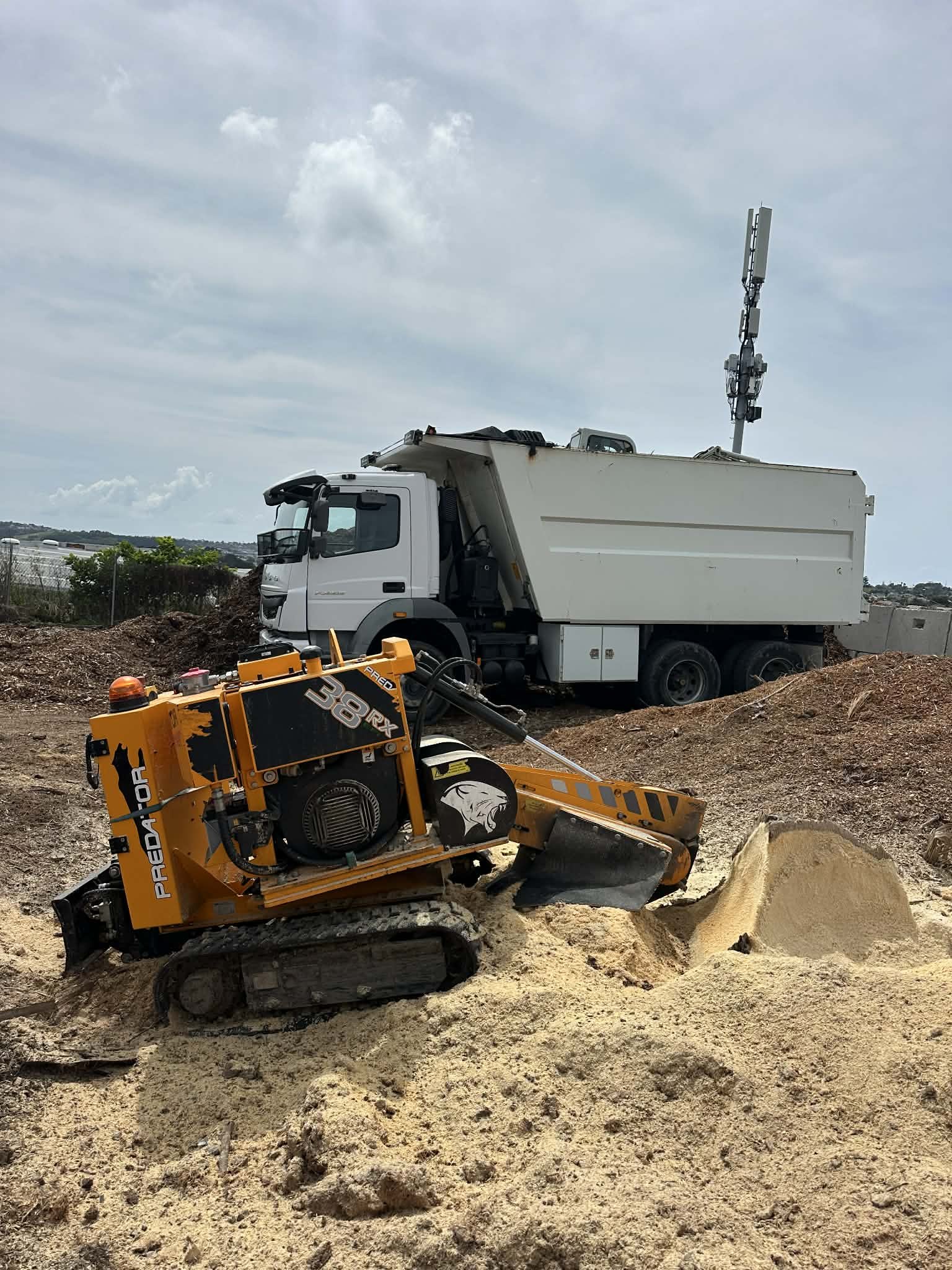 Construction site with a small orange stump grinder working on a mound of sand in the foreground and a white utility truck with antennas in the background under a cloudy sky.