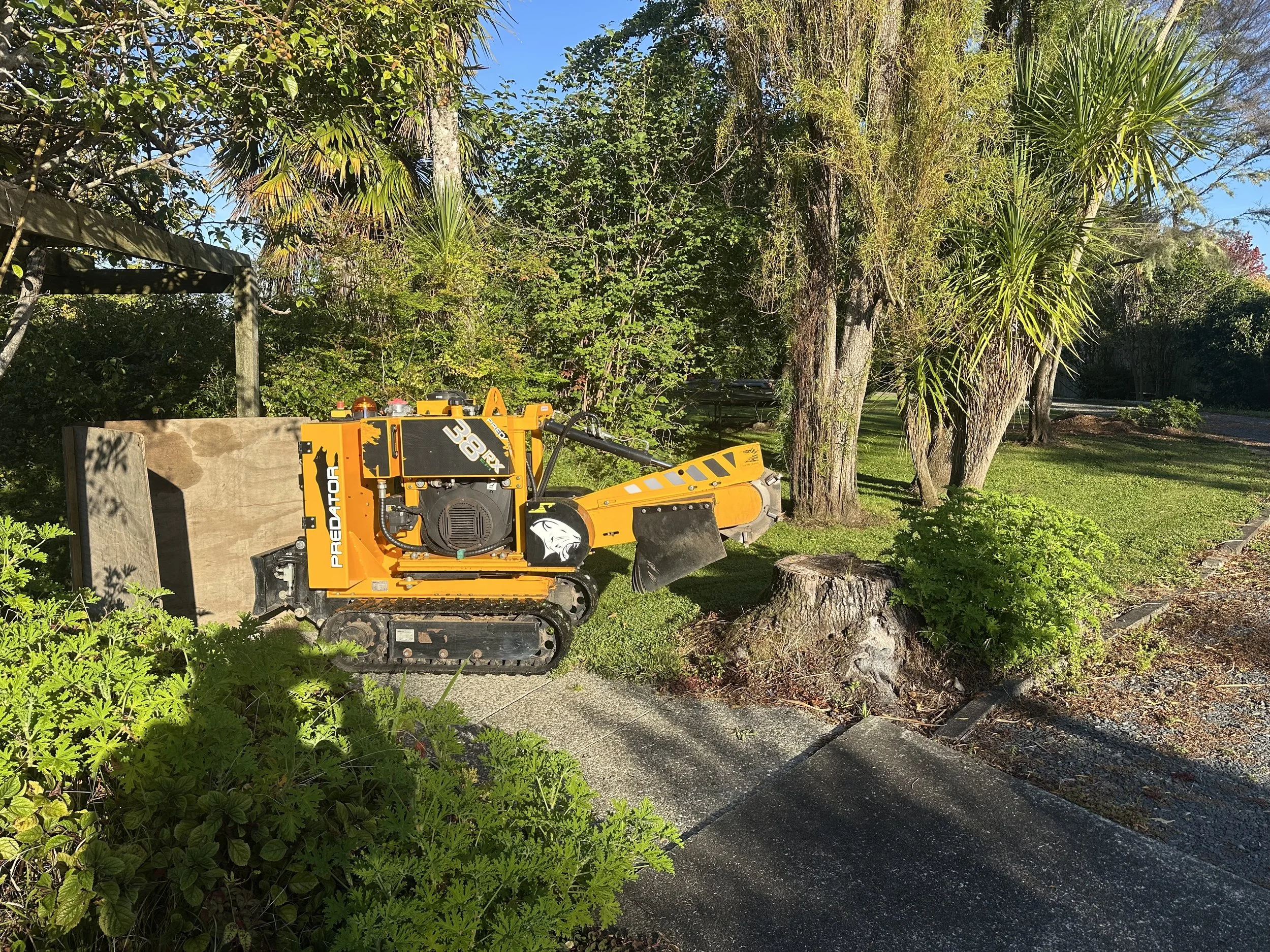 A small orange and black tracked stump grinder machine resting on the edge of a grassy lawn next to a tree stump, with trees and bushes in the background on a sunny day.