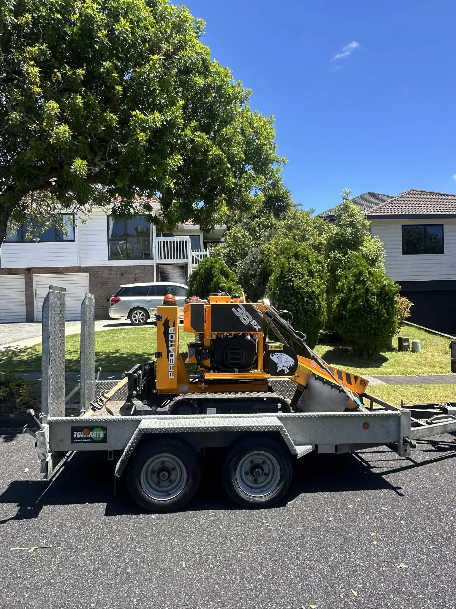 A small orange and black stump grinder named Predator on a trailer parked on the street in a residential neighborhood with houses, cars, green trees, and a clear blue sky in the background.