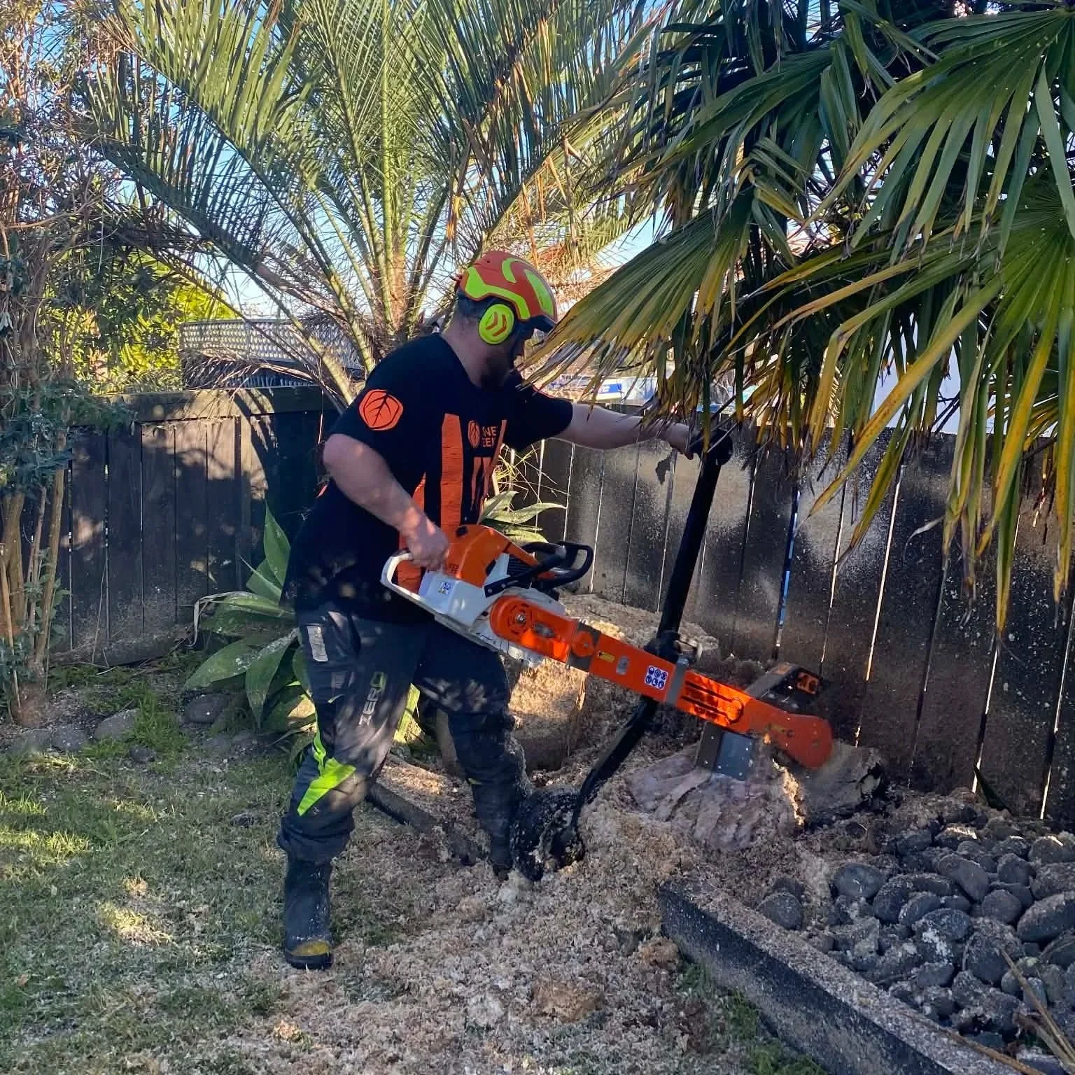A person wearing safety gear, including a helmet and protective clothing, is using an alpine handheld stump grinder to remove a tree stump in a backyard with a wooden fence, plants, and trees.