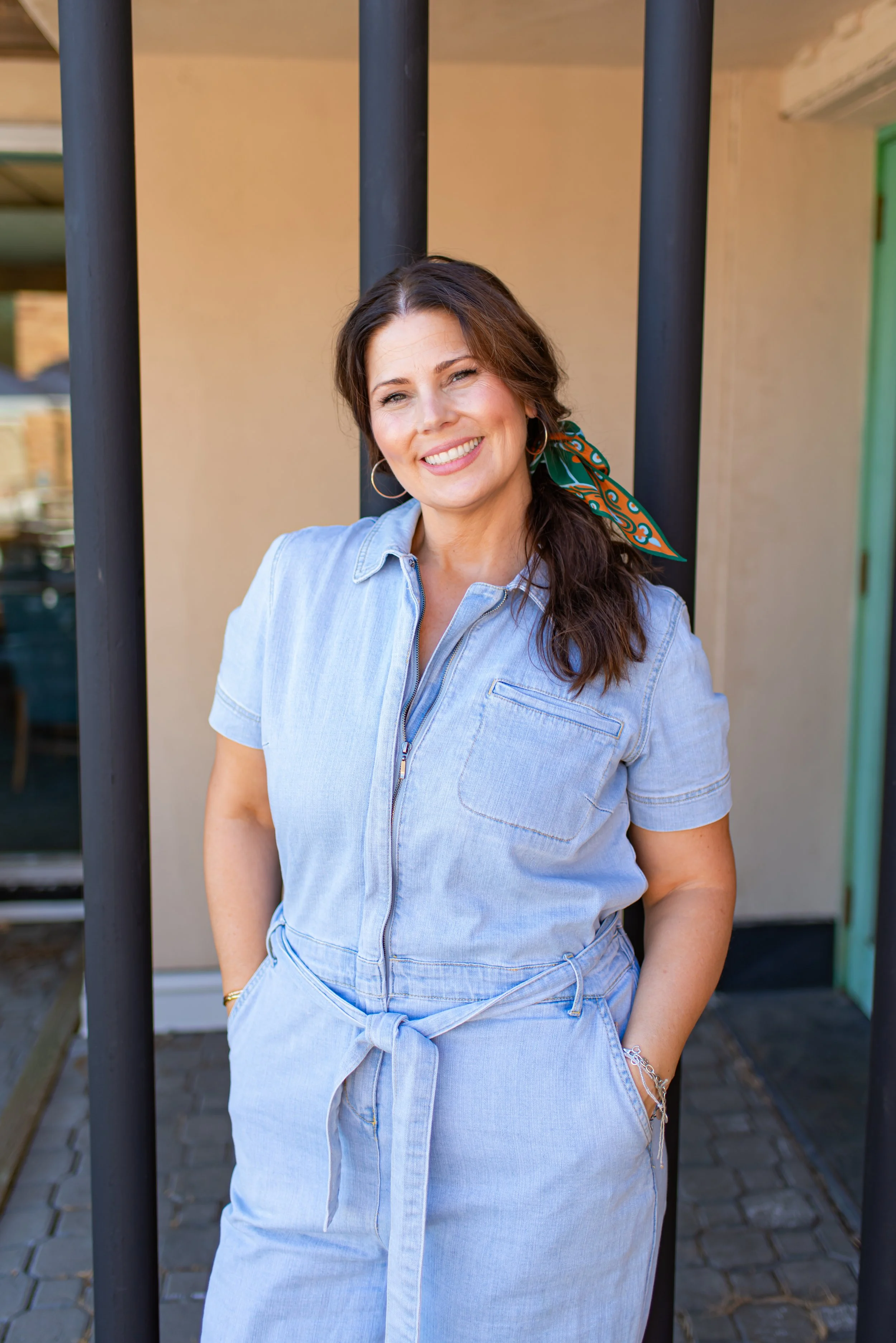 Young woman wearing light blue denim jumpsuit, standing outdoors near a black metal railing, smiling, with a bandana tied in her hair.