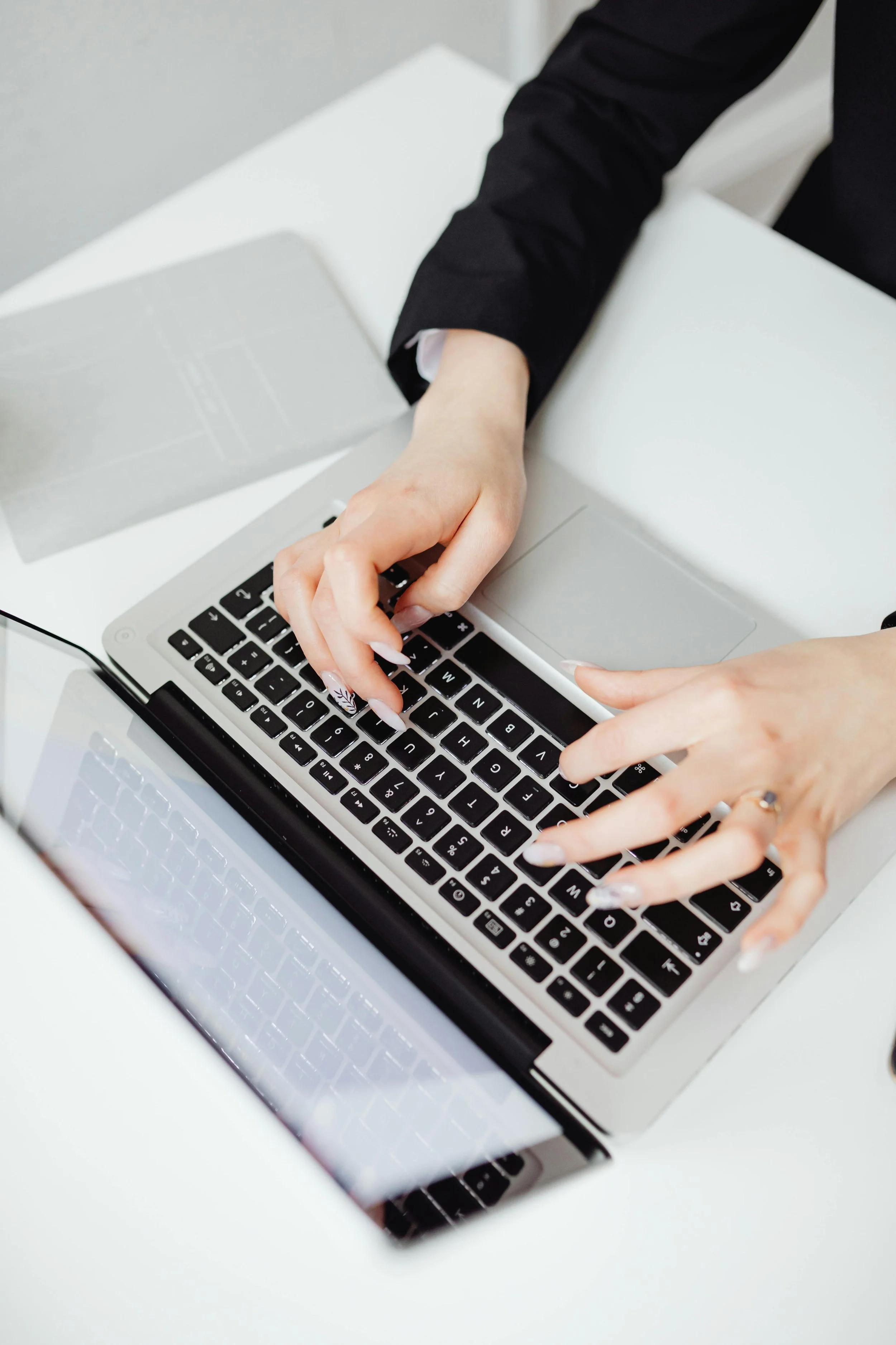 Person typing on a silver laptop keyboard at a white desk, dressed in a black sleeve shirt.