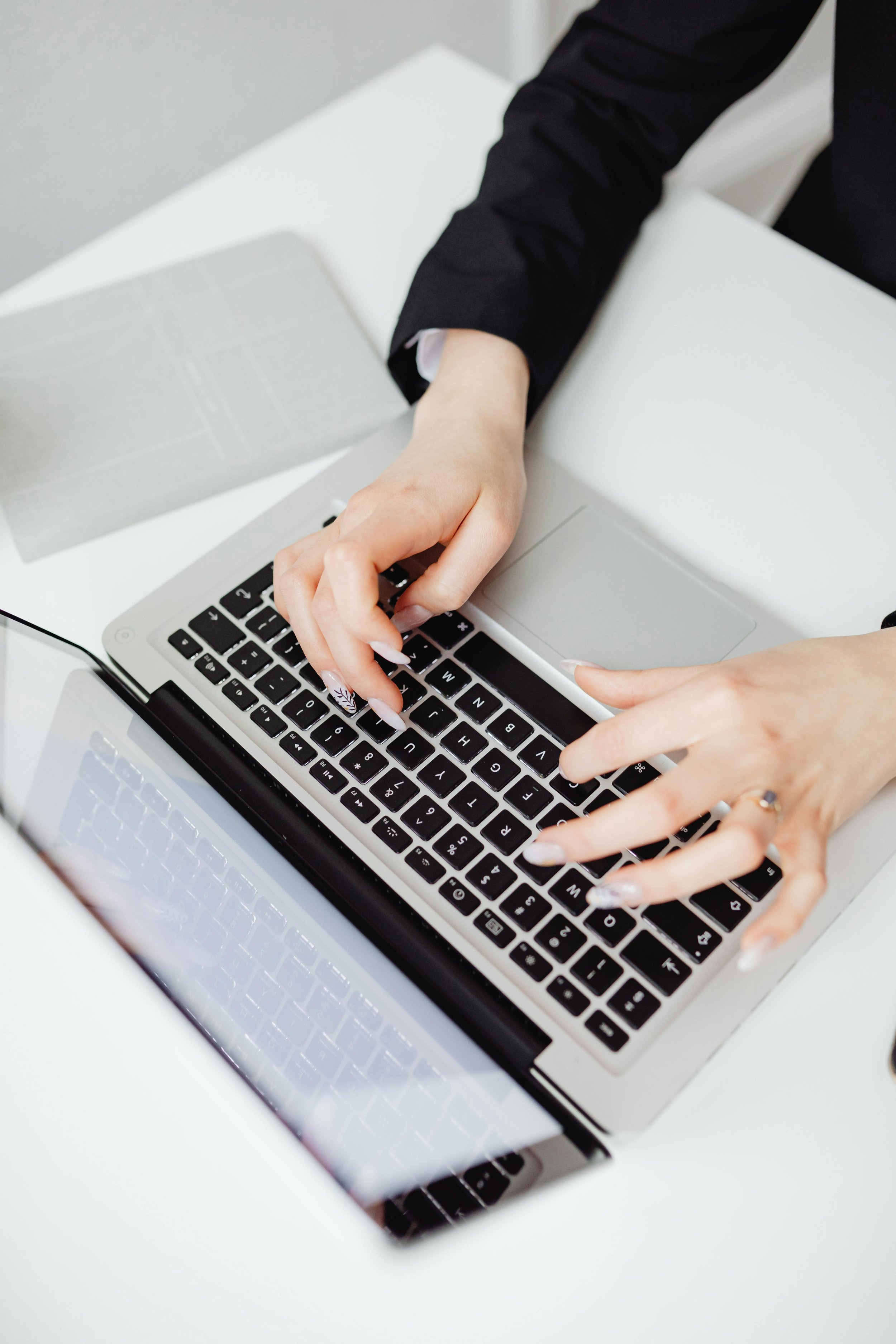 Person typing on a silver laptop at a white desk, wearing a black blazer.