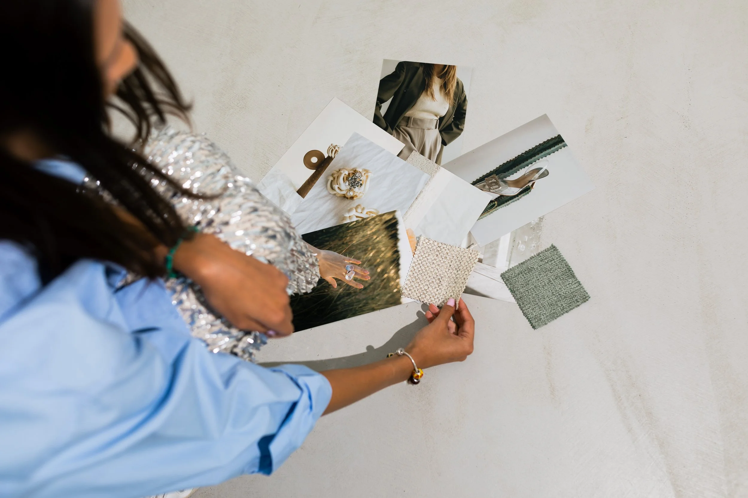 A woman arranging fabric and photo samples on a white surface, surrounded by various textiles and photographs, with her left hand holding a beige fabric sample.