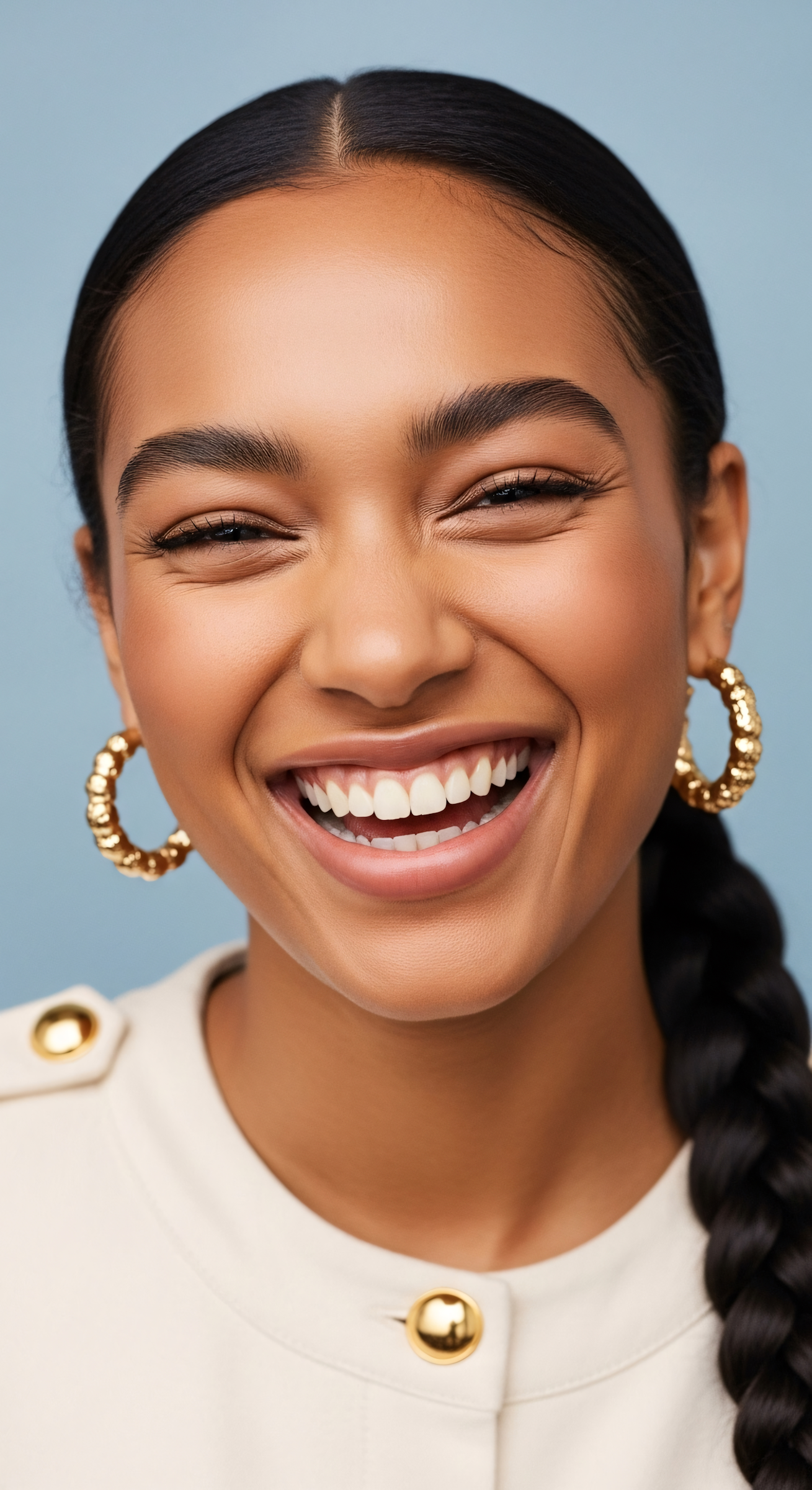 Close-up of a smiling woman with dark hair tied in a braid, wearing large gold hoop earrings and a cream-colored top with gold buttons, against a plain blue background.