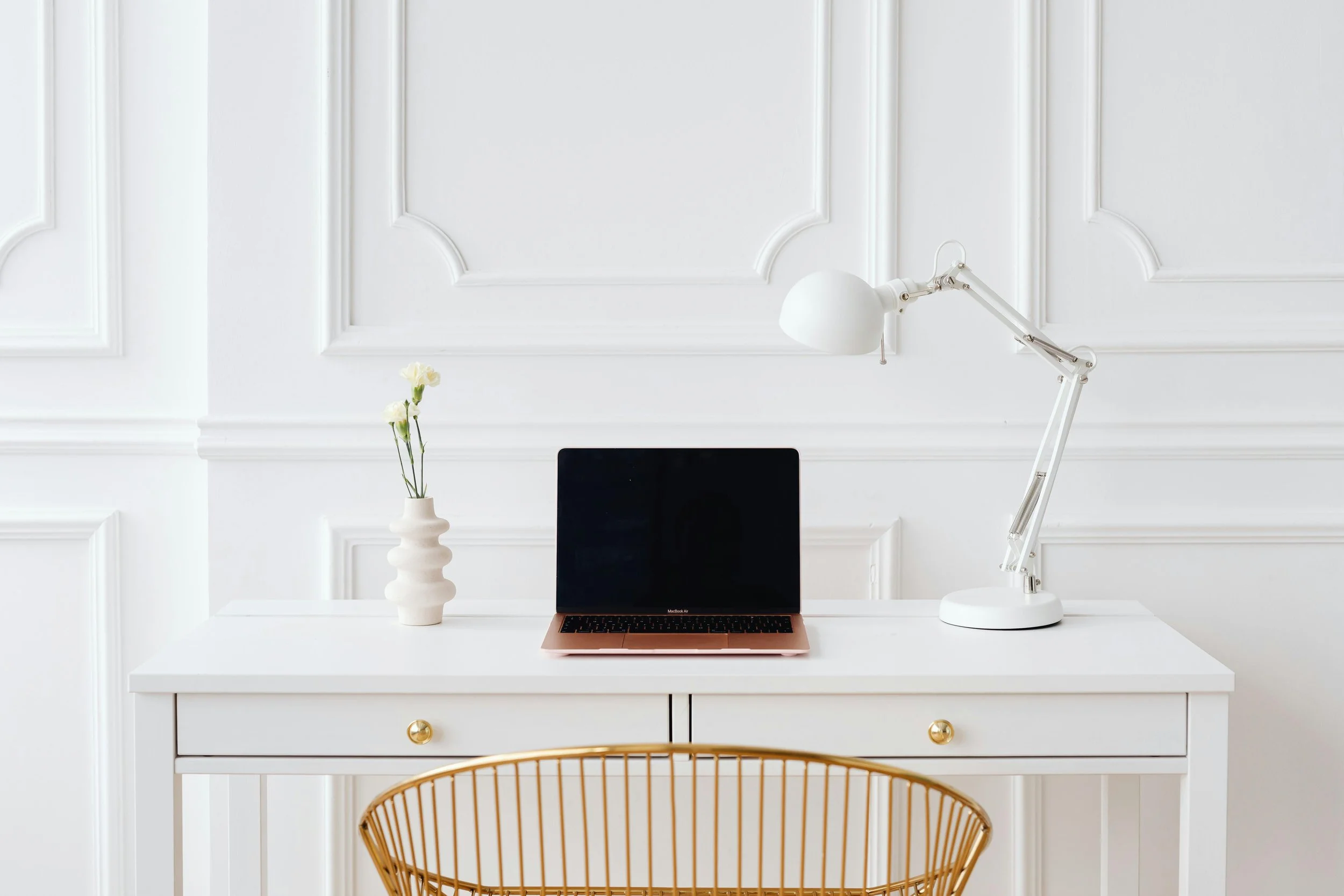 A white desk against a white wall with decorative molding, featuring a vase with white flowers, a closed rose gold laptop, and a white adjustable desk lamp, with a gold wire chair in front.