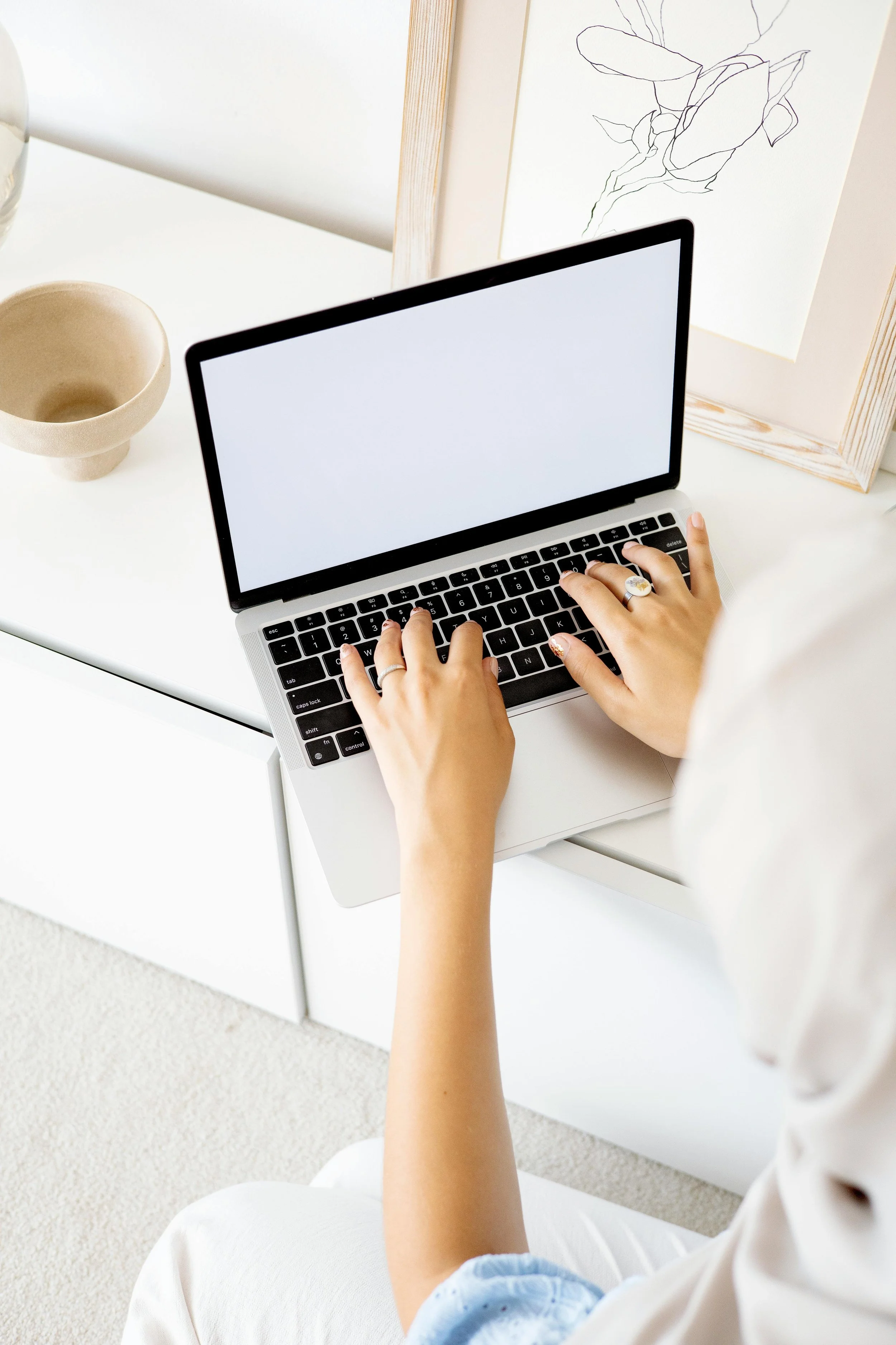 A person using a laptop at a white desk, with a beige cup and a framed line art drawing of a flower on the wall.