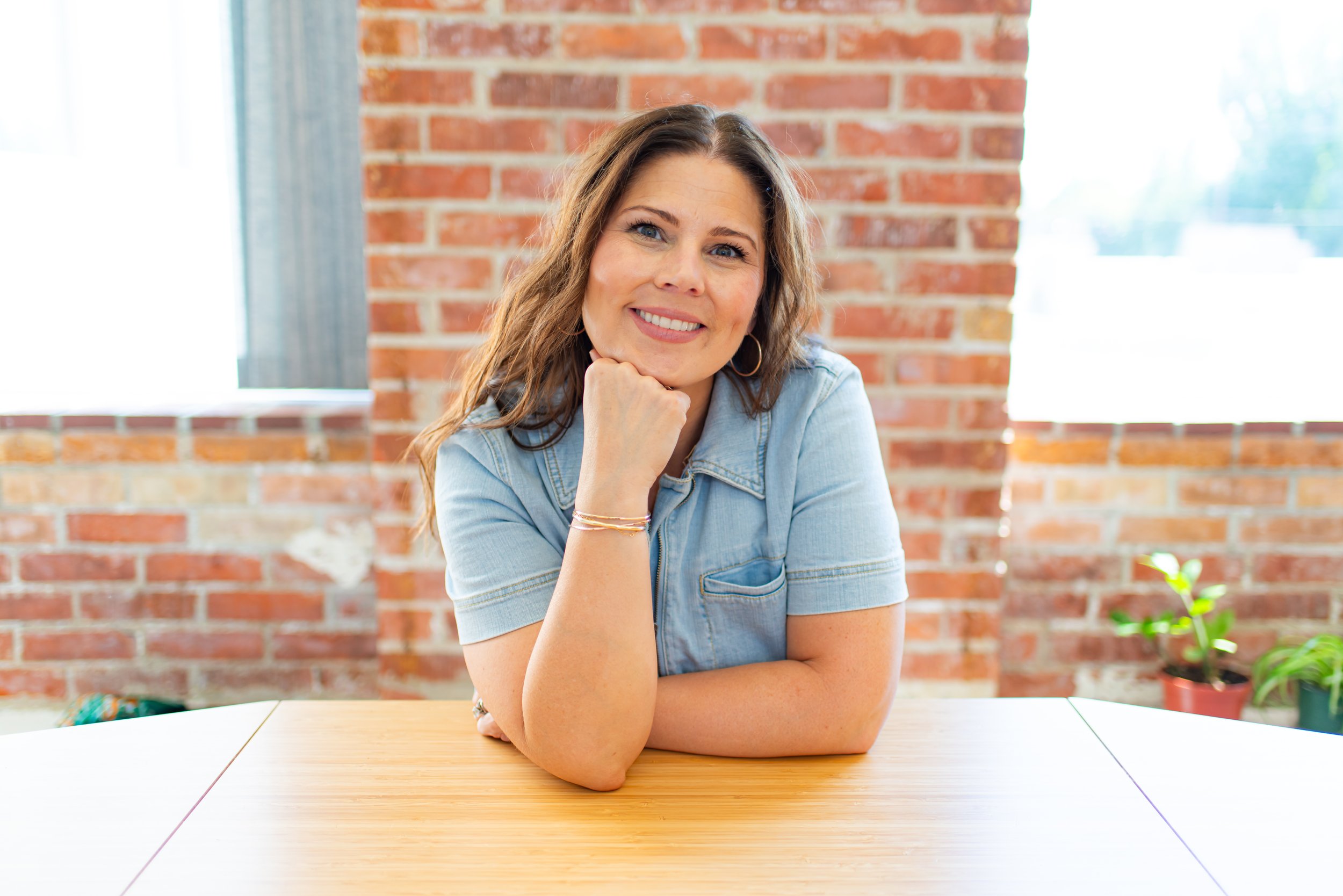 A woman with shoulder-length brown hair, wearing a light denim shirt and gold hoop earrings, is sitting at a wooden table with her chin resting on her hand, smiling at the camera. Behind her is a brick wall with windows and some potted plants.