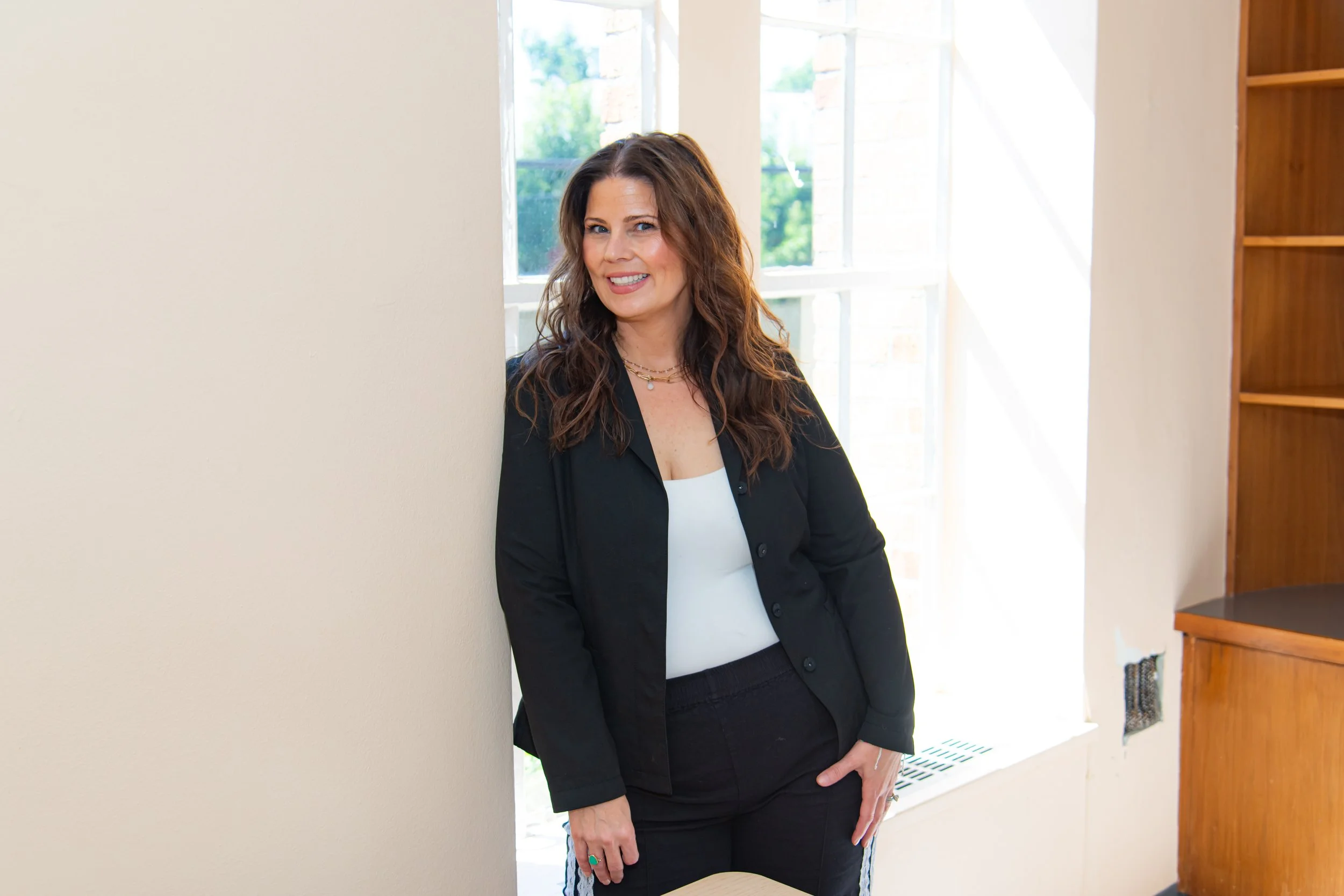 A woman with long wavy brown hair standing indoors near a wall and large window, smiling at the camera, wearing a black blazer over a white top, with jewelry, in a well-lit room with wooden furniture.