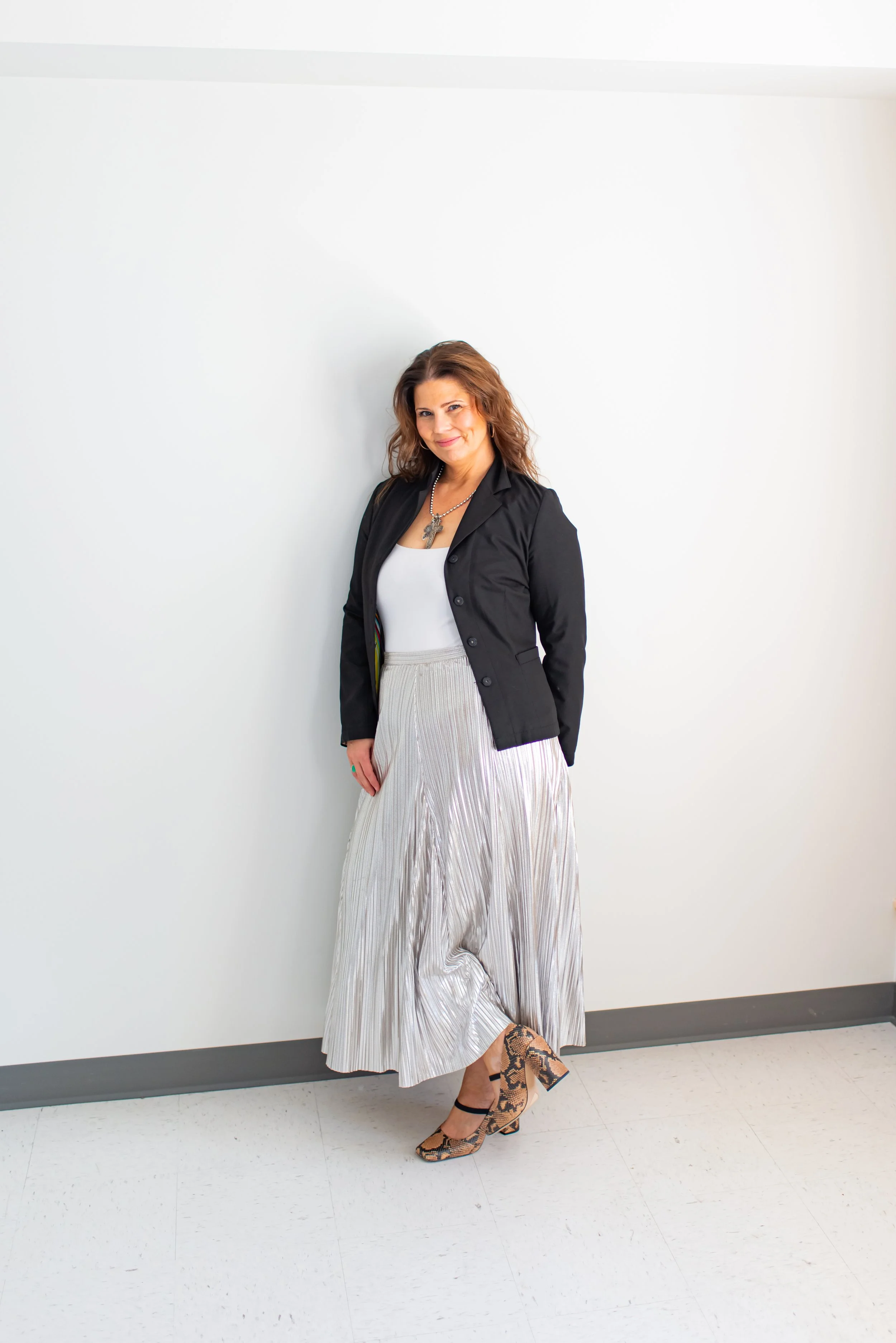 A woman with brown, shoulder-length hair wearing a black blazer, white top, long pleated shiny skirt, and snakeskin heels, standing against a white wall and smiling.