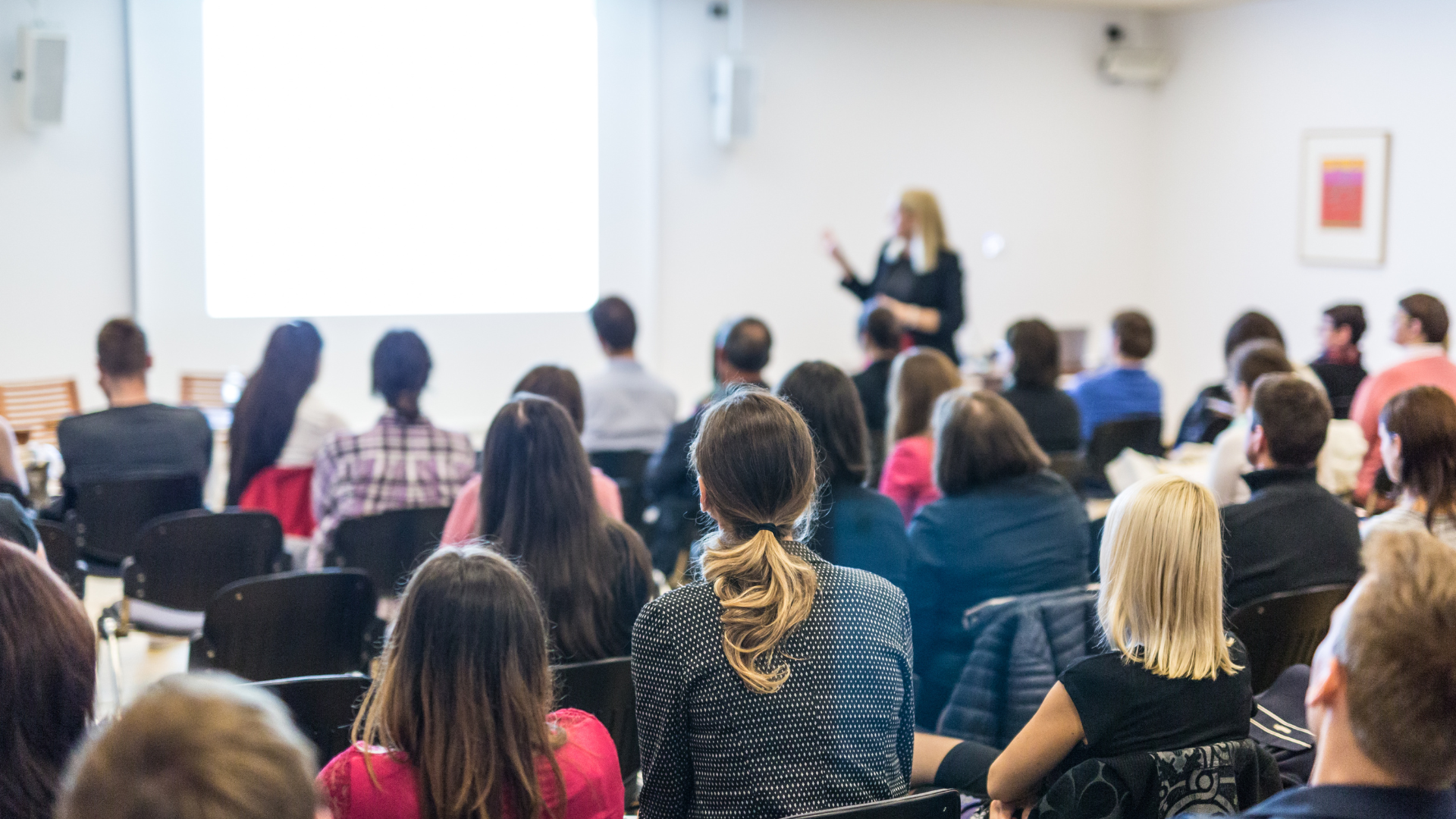 A woman giving a presentation to an audience in a conference room. The audience is seated and facing a large screen, with some taking notes.