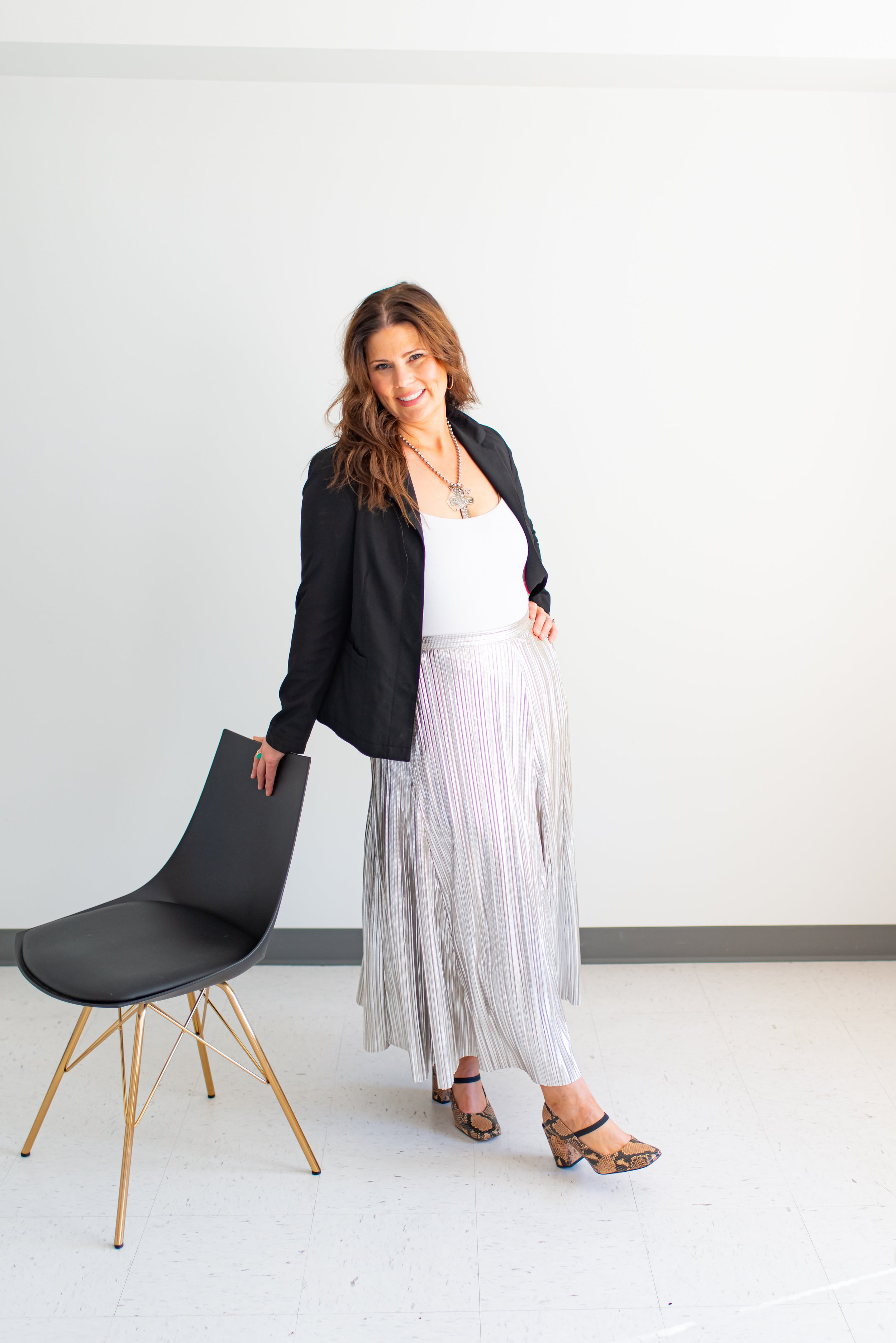 A woman with long brown hair, wearing a black blazer, white top, beige striped maxi skirt, and snakeskin heels, standing next to a black chair with gold legs against a plain white wall, smiling at the camera.