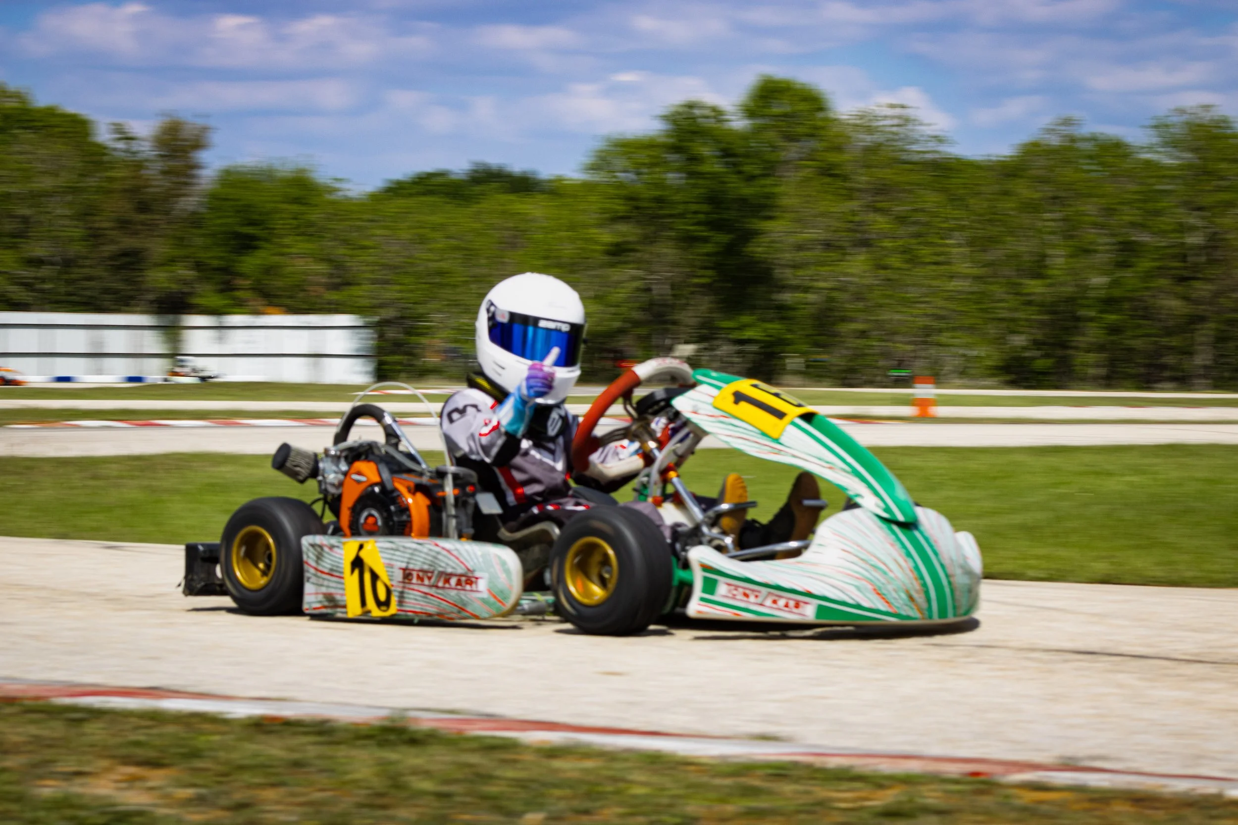 Go-kart racing on a track with a driver wearing a white helmet and racing suit, giving a thumbs-up gesture.
