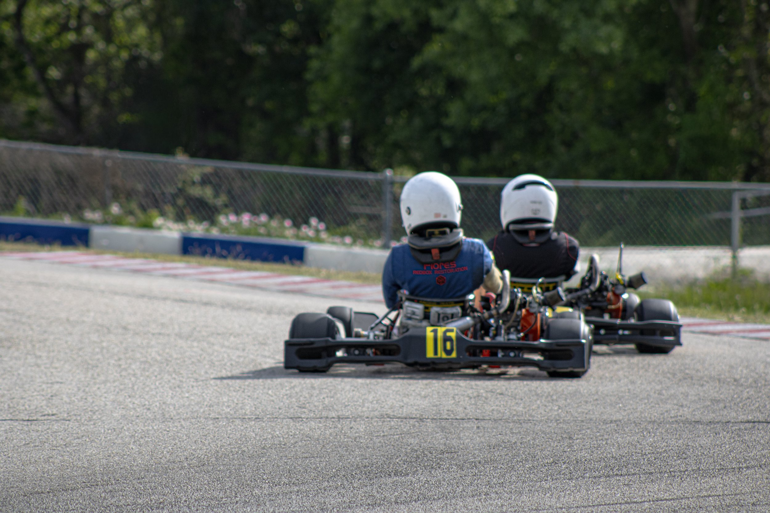 Two people racing go-karts on an outdoor track, both wearing white helmets and dark racing suits, with the kart number 16 visible in the foreground.