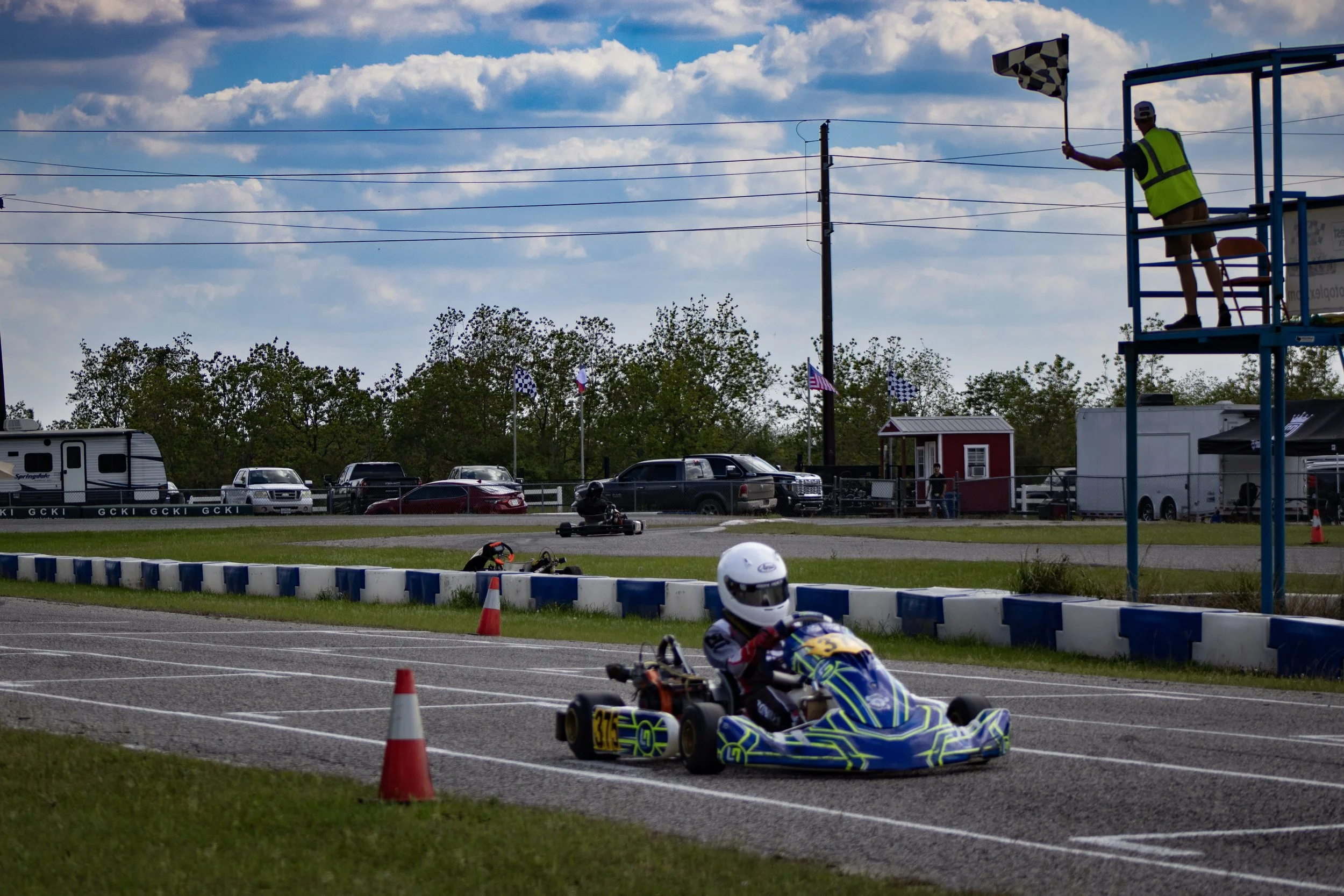 Go-kart racer in a blue and yellow kart on a track, with a racetrack official on a raised platform waving a checkered flag, and cars parked in the background.