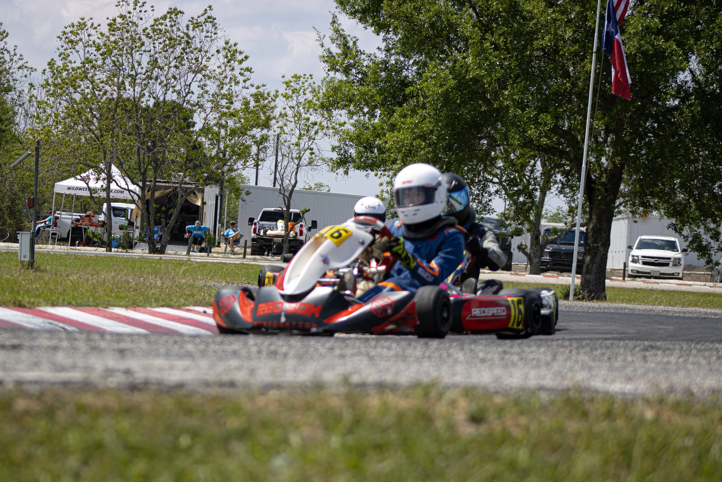 Two race car drivers in helmets and racing suits navigate a turn on a race track in go-karts, with spectators and parked vehicles in the background under a partly cloudy sky.