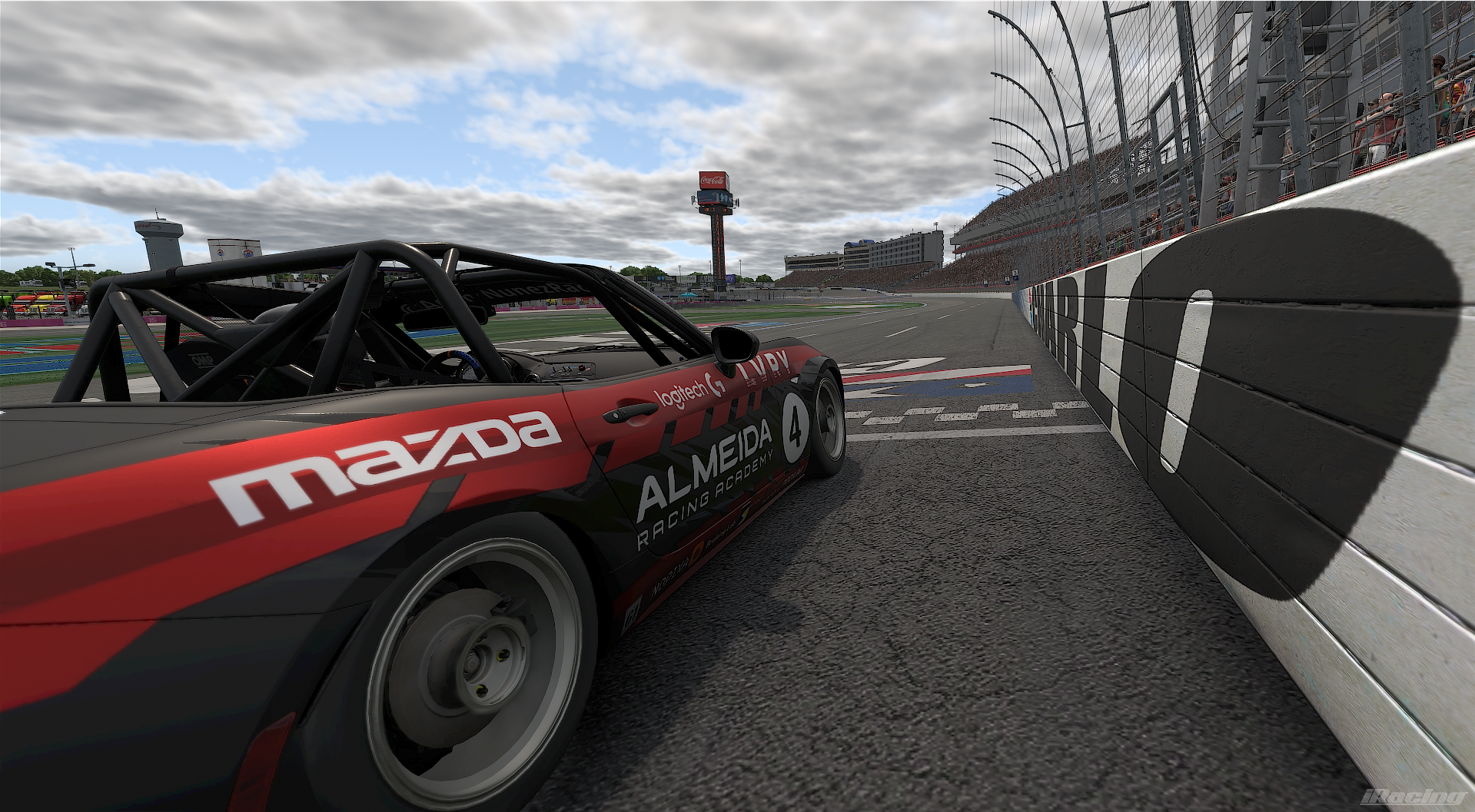 A racing car at the finish line of a race track, with a large crowd and grandstands in the background, and a cloudy sky overhead.