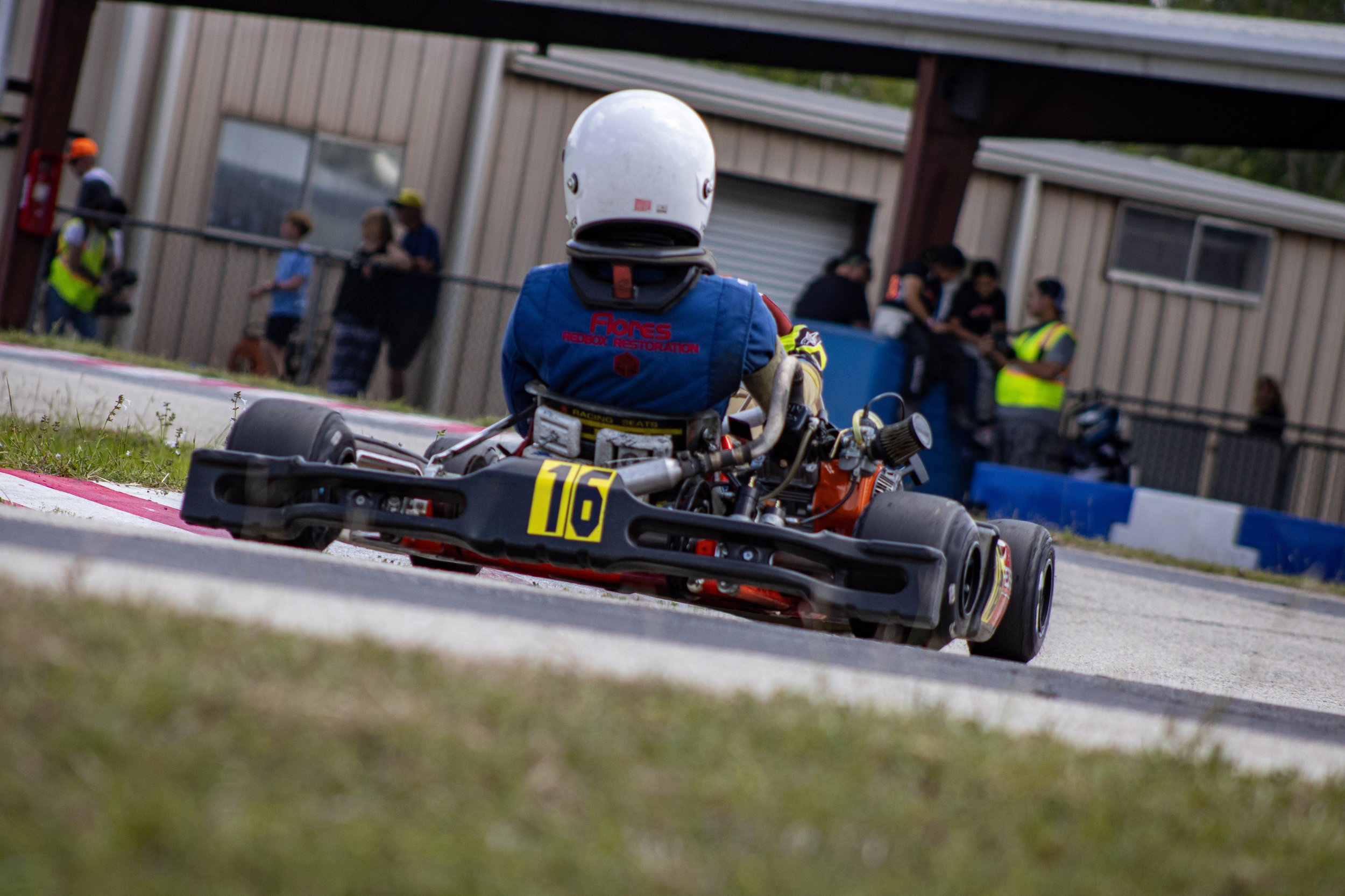 A go-kart racer wearing a white helmet and blue racing suit with 'Flores' logo, driving a go-kart with the number 16 on the front, on a race track with spectators and officials in the background.