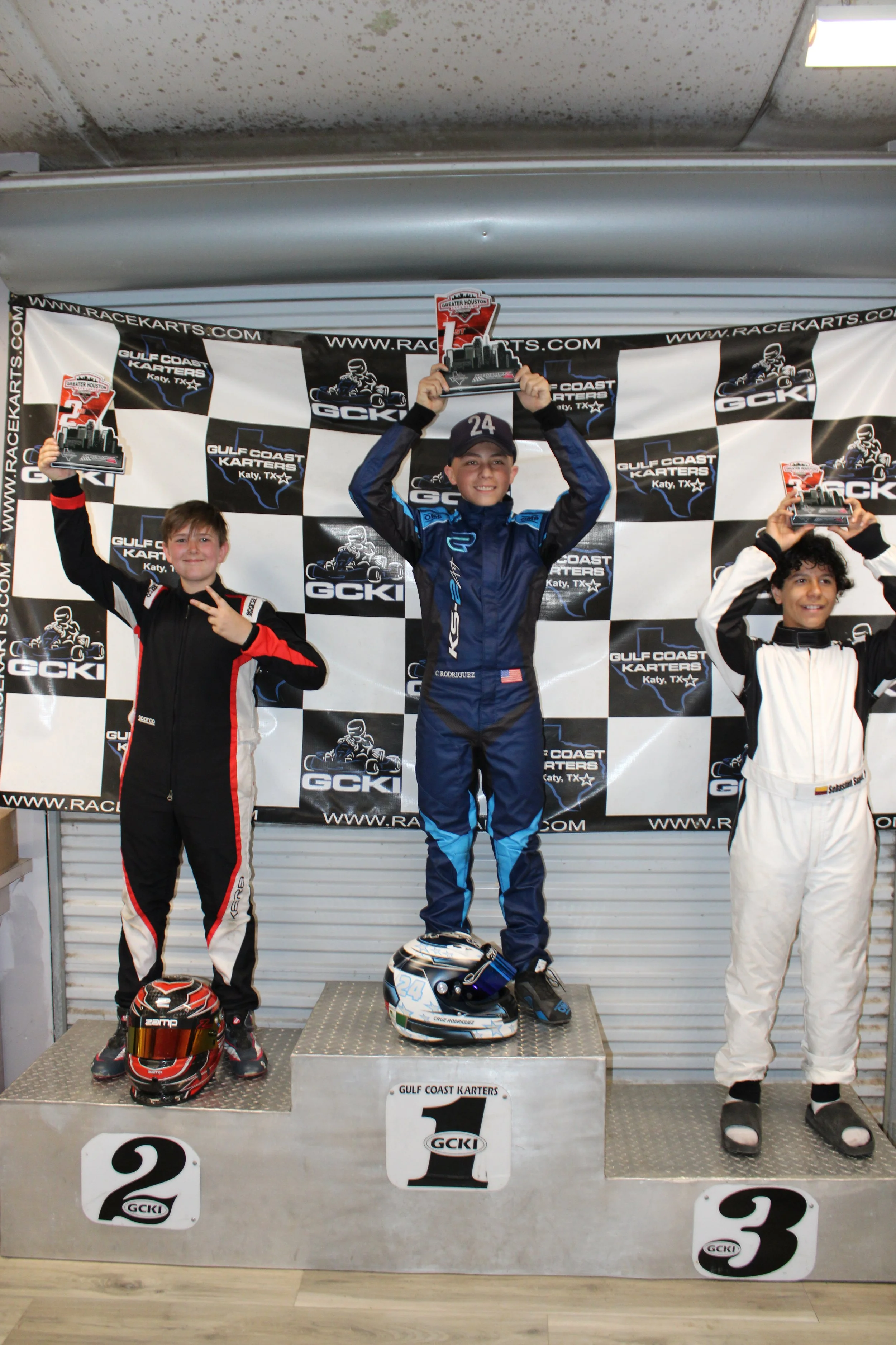 Three young go-kart racers standing on a winners' podium holding trophies, with race helmets on the ground, in front of a backdrop for Gulf Coast Karts in Katy, Texas.