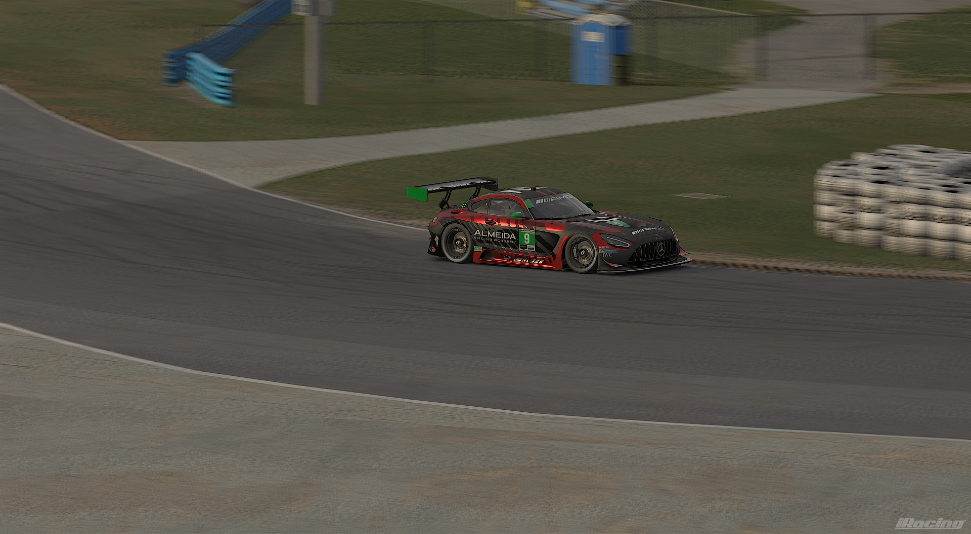 A black and red race car with number 9 on its side is on a race track, heading into a sharp turn. The track has tire barriers on the side and a grassy area with a slide and a restroom in the background.