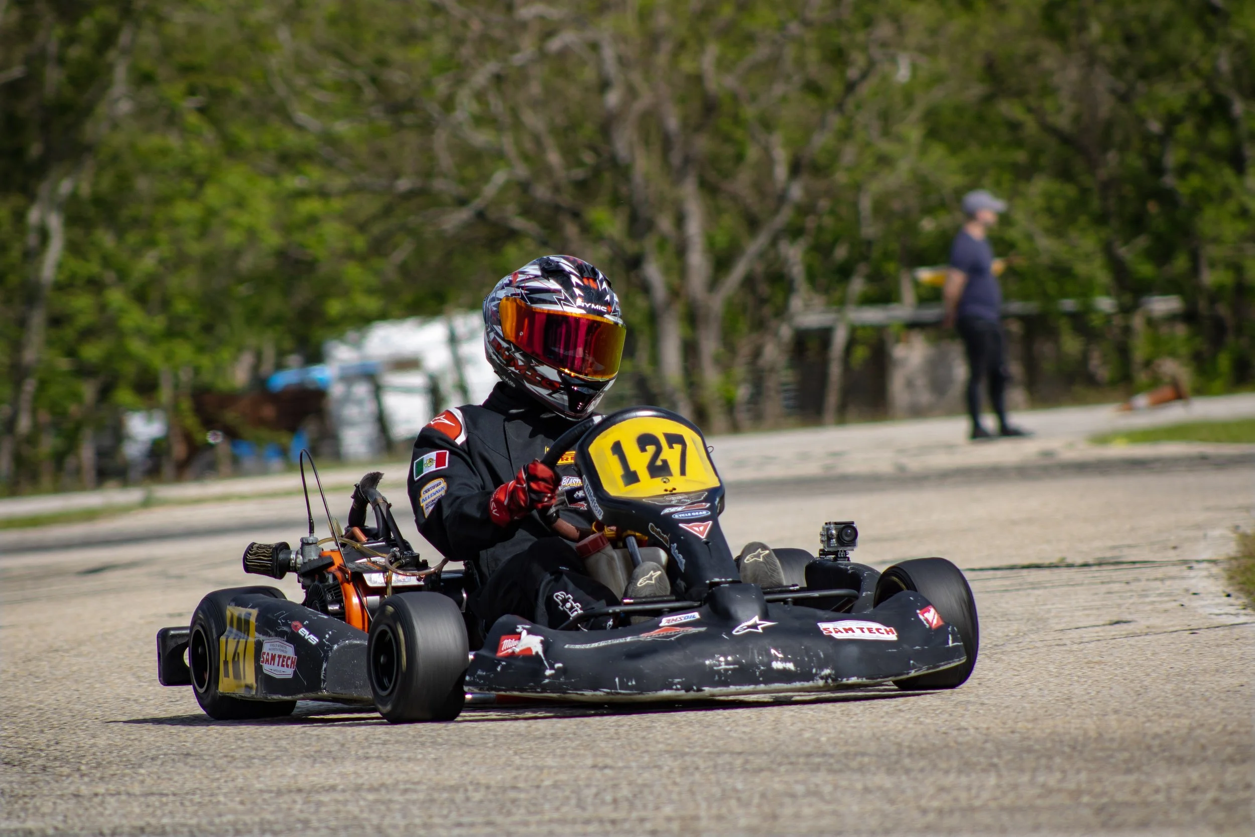 Go-kart racer wearing a black racing suit and helmet, number 127, driving on a track with trees in the background.