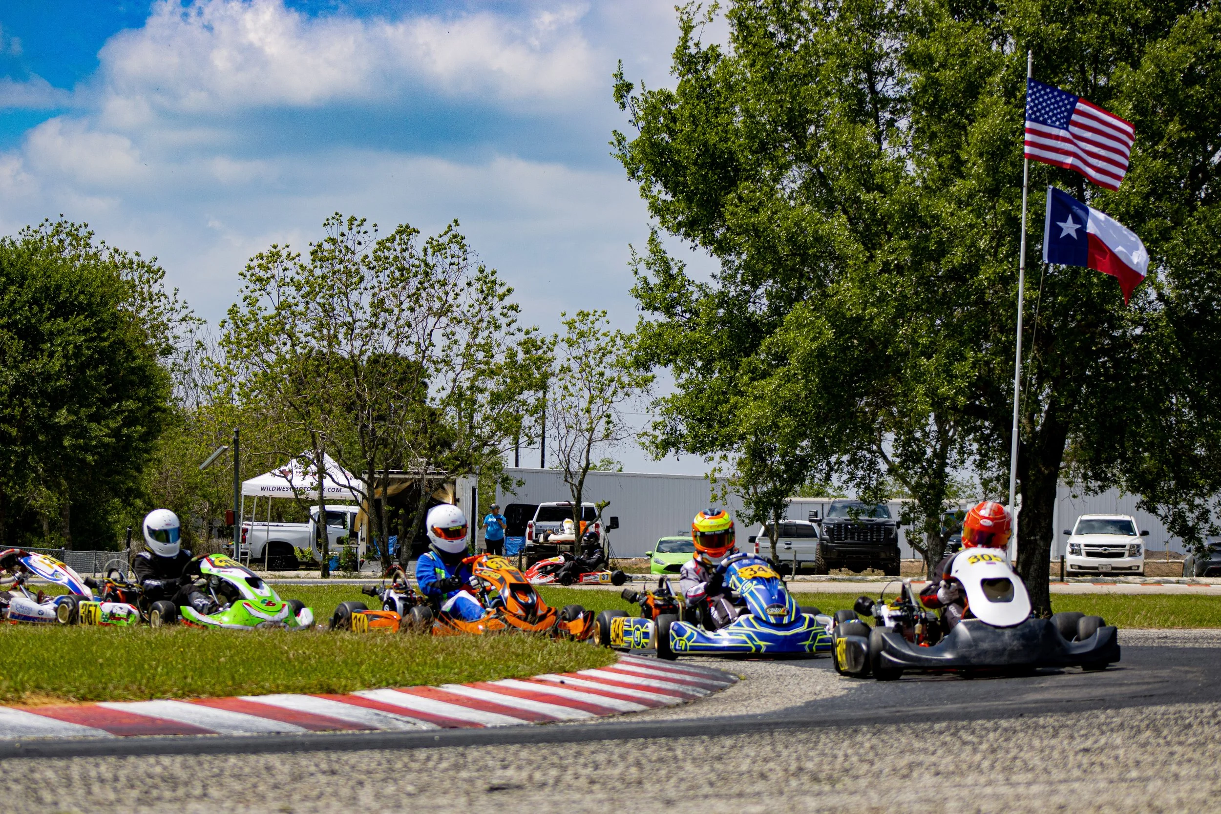 Go-kart racers on a track with trees, vehicles, and flags in the background.