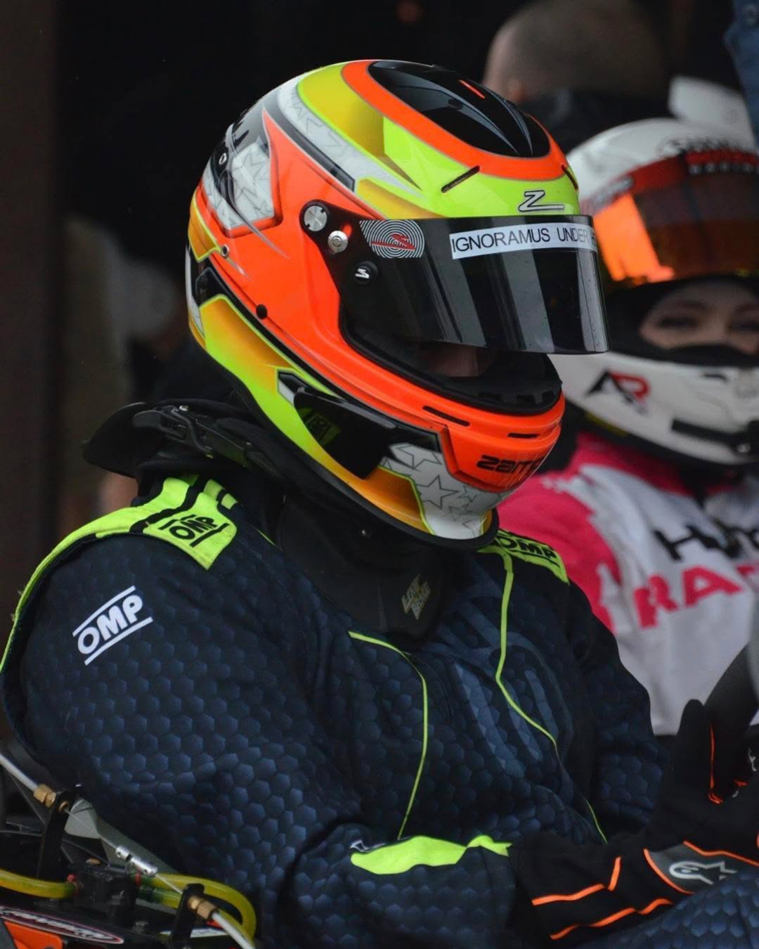 Close-up of a race car driver wearing a bright orange, yellow, and black helmet and racing suit with neon yellow accents, seated in a race car garage.