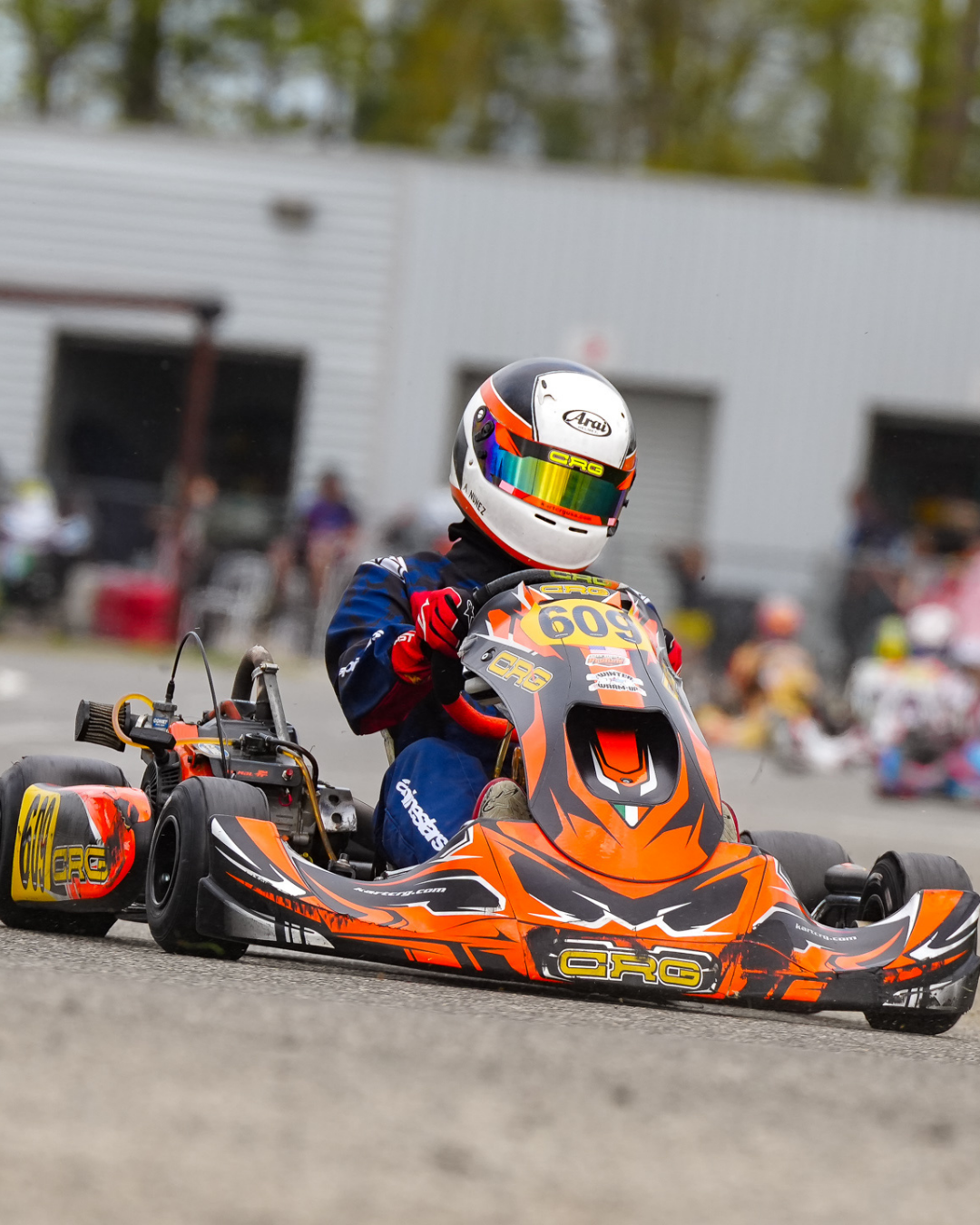 Go-kart racer wearing helmet and racing suit sitting in a bright orange and black go-kart on track, with a blurry background of garage and other racers.