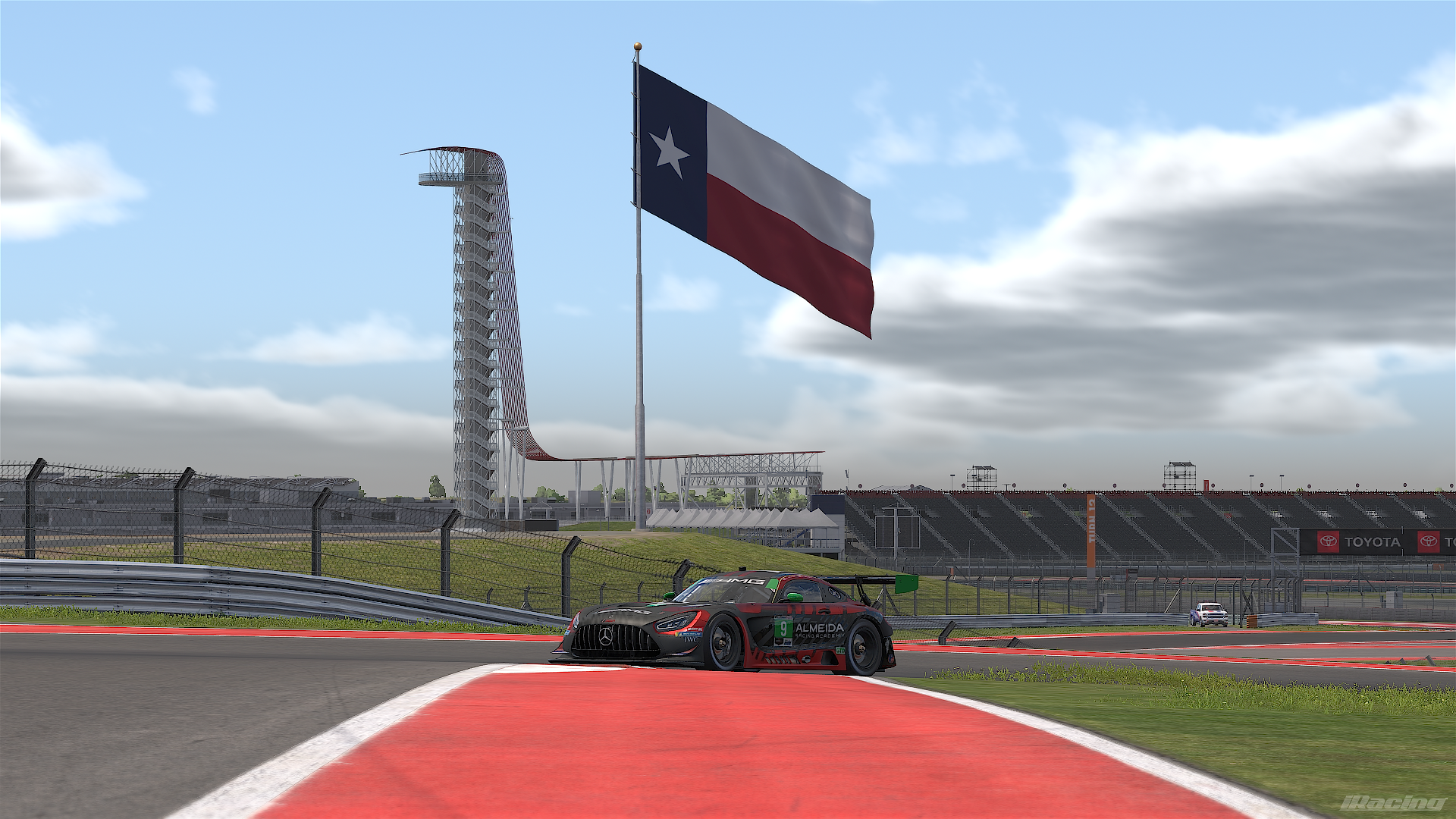 A race car on a track with a large Texas flag flying in the background. The scene is set at a racing circuit with grandstands and a tower visible under a partly cloudy sky.
