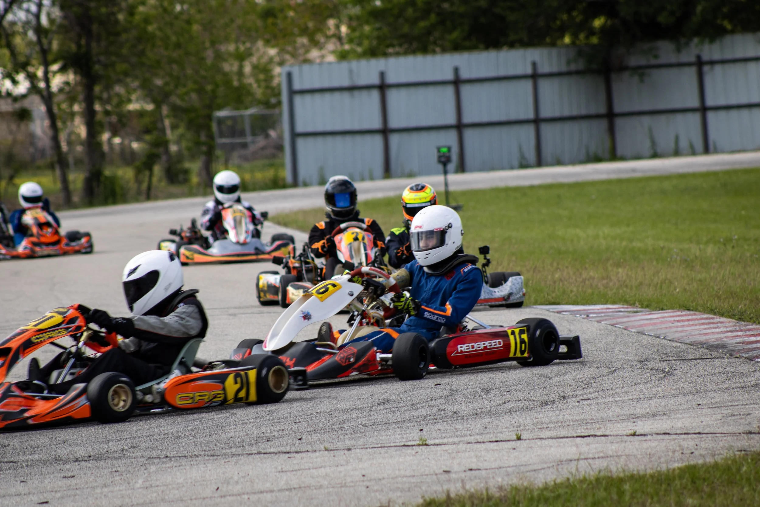 Children racing on go-karts on an outdoor track, all wearing helmets and protective gear.