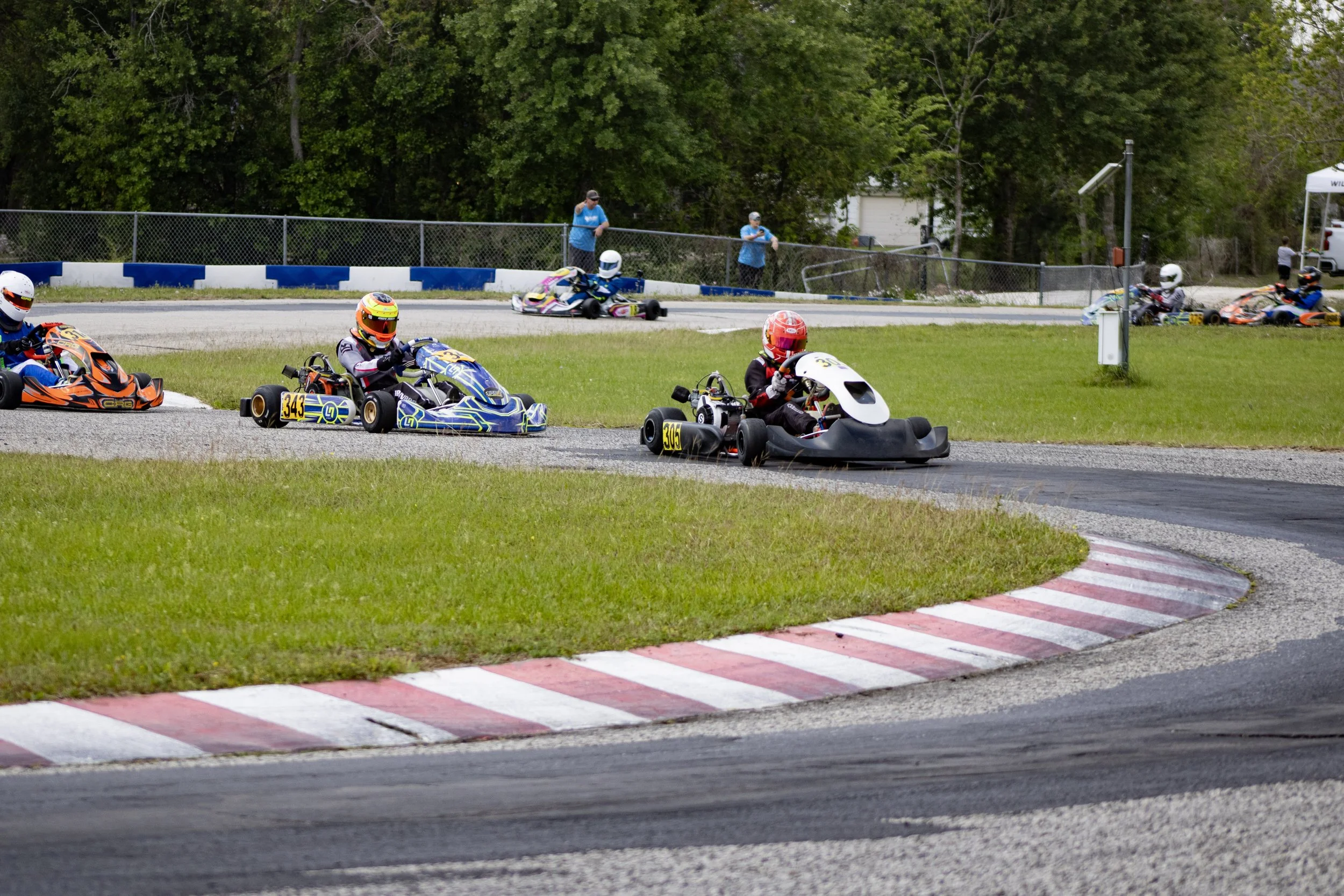 Go-kart race with multiple racers navigating a turn on a track surrounded by grass and trees, with some people watching in the background.