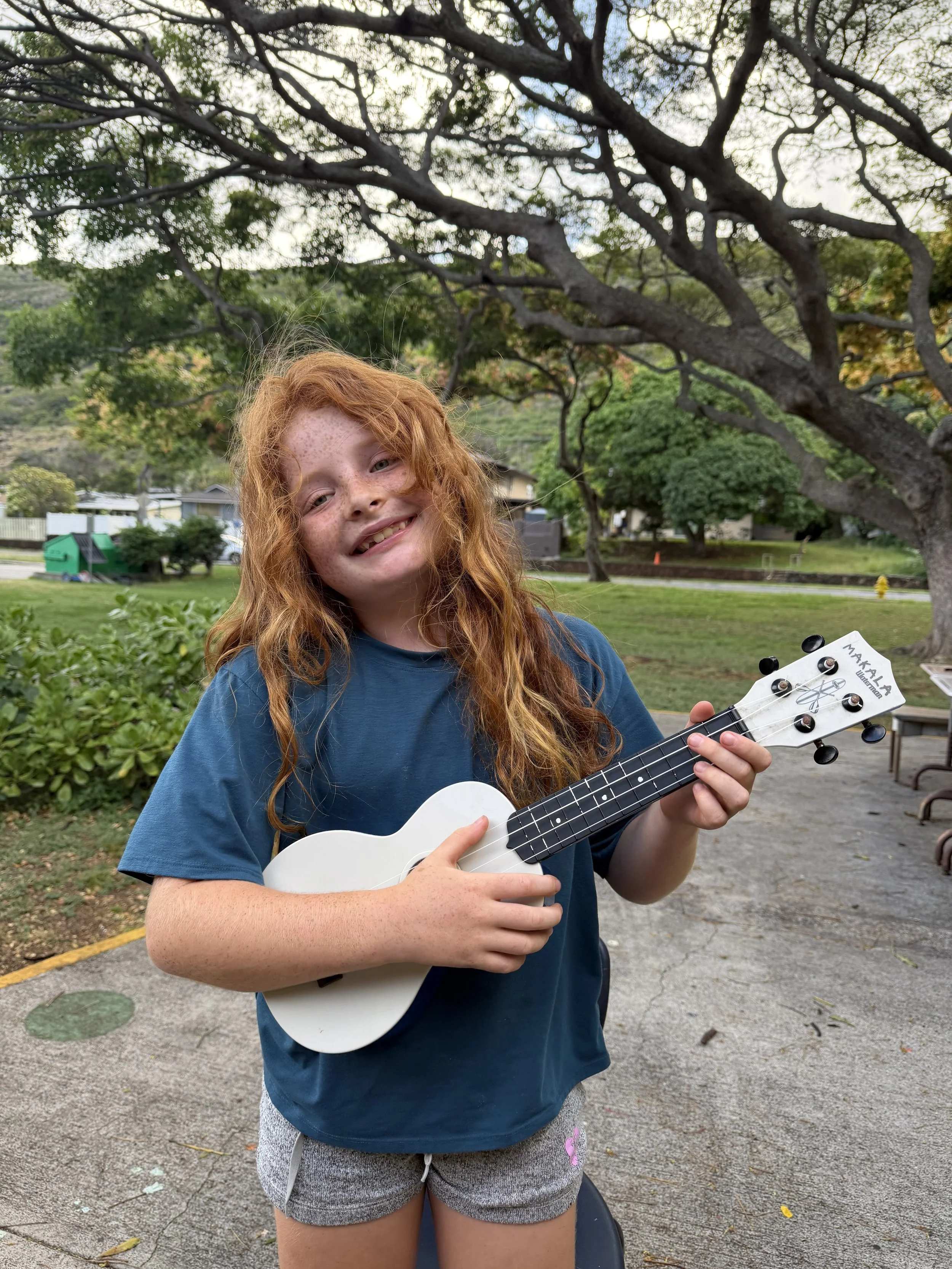 A young girl with red hair and freckles smiling while holding a small white ukulele in an outdoor park setting with trees and a building in the background.