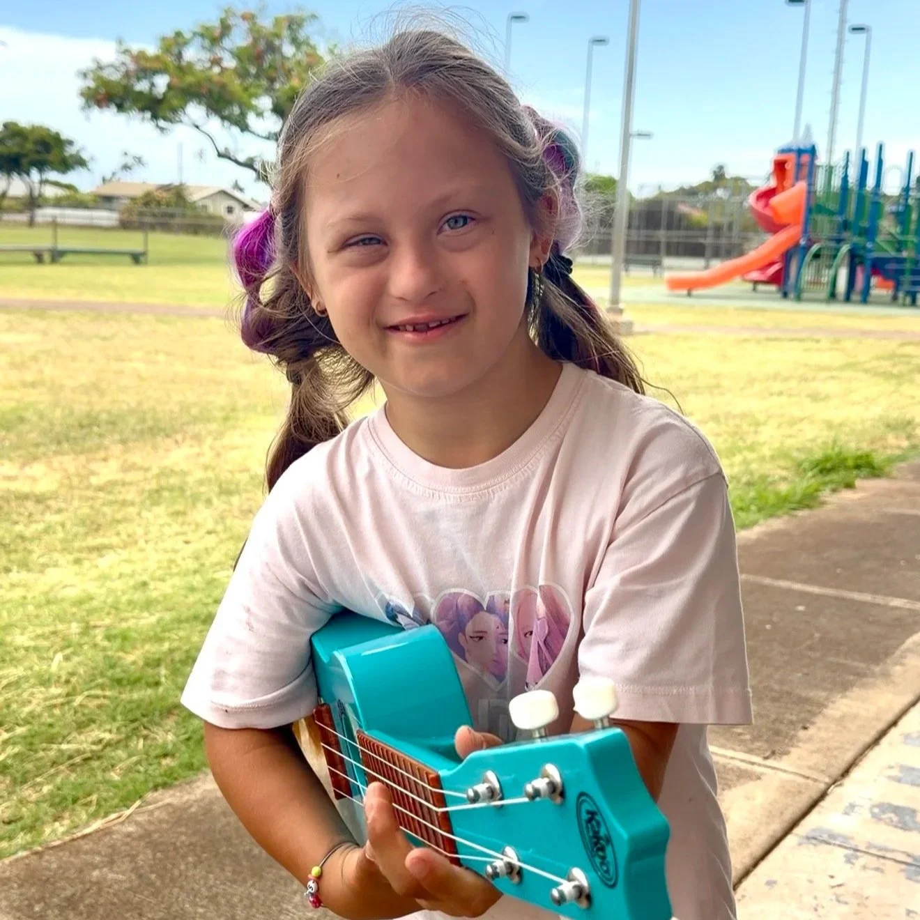 A young girl with braided hair accented with purple and pink streaks, holding a blue ukulele, standing outdoors at a park with playground equipment and trees in the background.