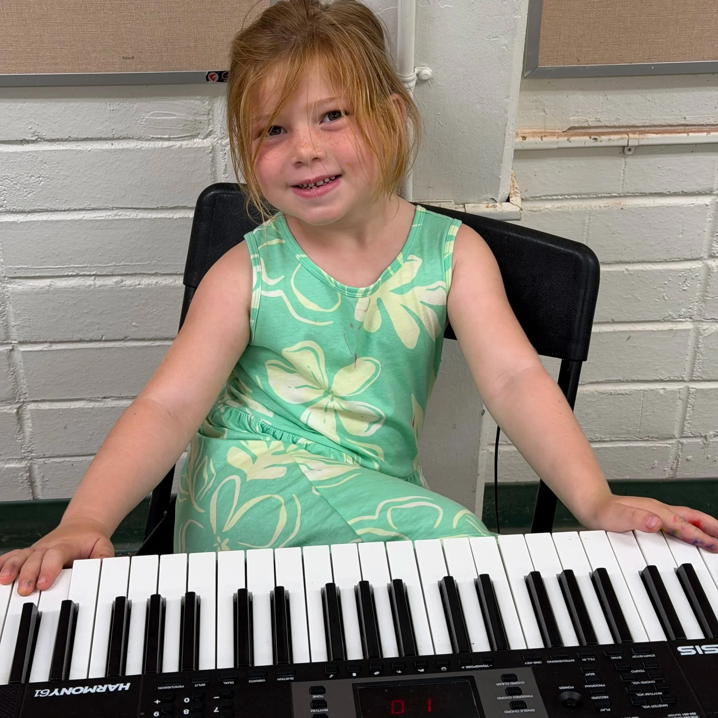 A young girl with red hair and freckles wearing a green sleeveless dress, sitting at a keyboard, smiling at the camera against a white brick wall background.