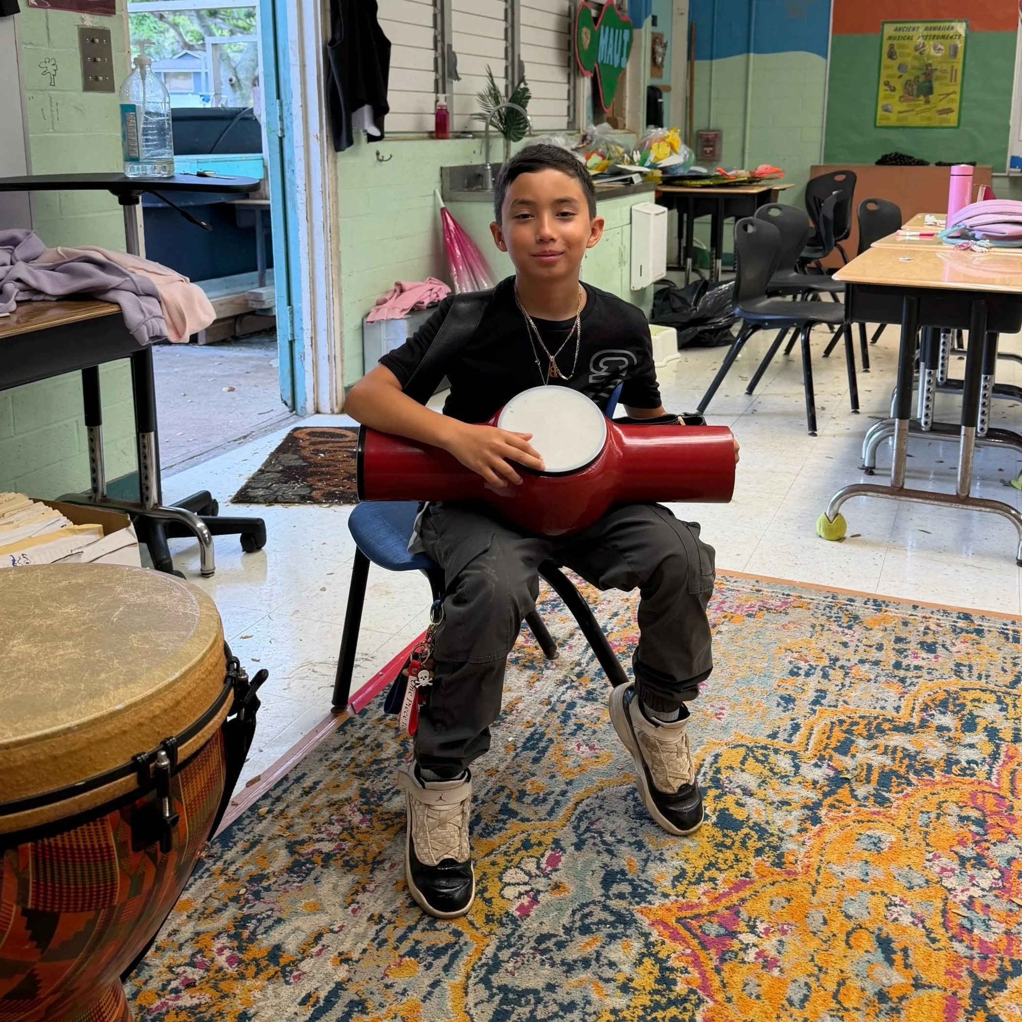 A young boy sitting on a chair in a classroom, holding a red drum with a white drumhead. There are musical instruments around him, including a large drum in the foreground and a tennis ball on the floor. The classroom has tables, chairs, and educatio