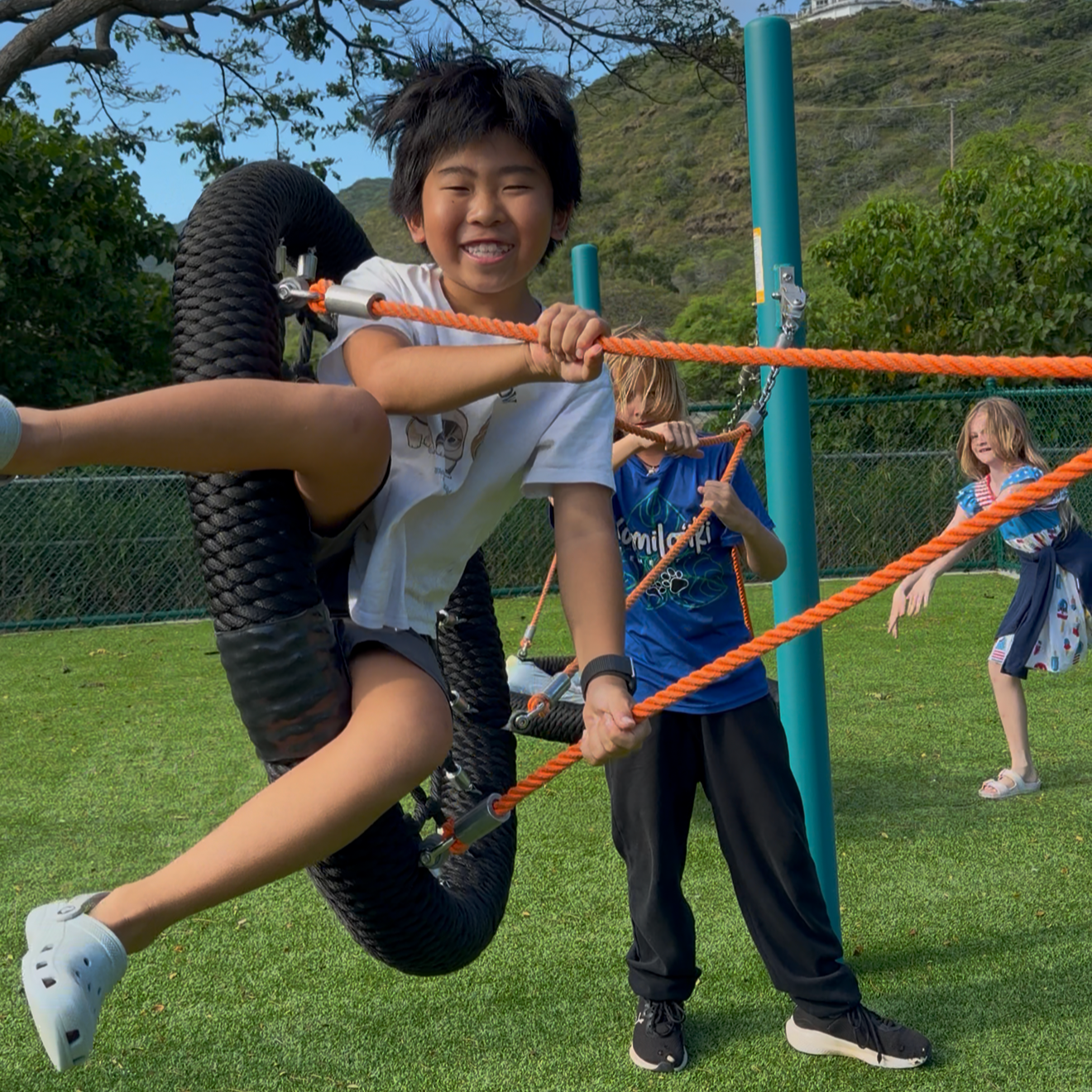 Smiling boy climbing on a rope ladder at a playground, with two girls in the background also playing, set outdoors with grass, trees, and a hillside.