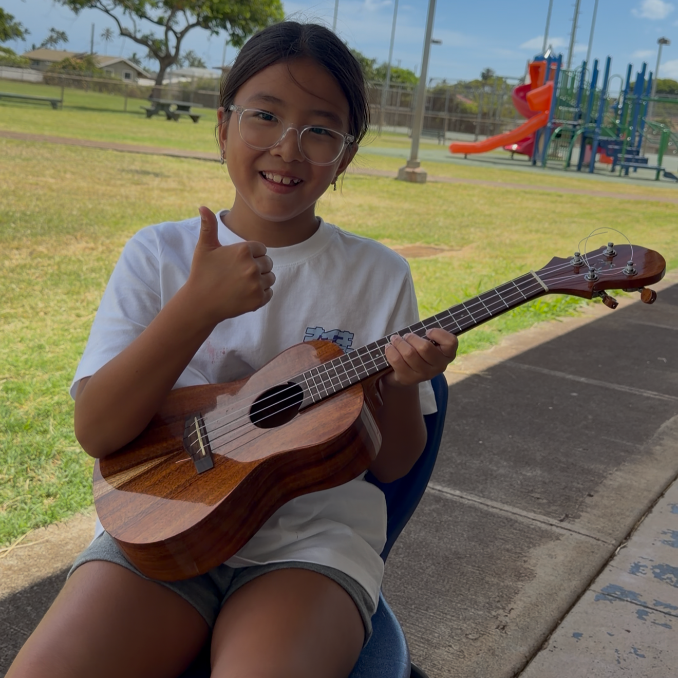A young girl wearing glasses and a white t-shirt is sitting outside, playing a small wooden ukulele. She is smiling and giving a thumbs-up gesture. Behind her, there is a grassy area, a playground with slides, and a clear sky with some clouds.