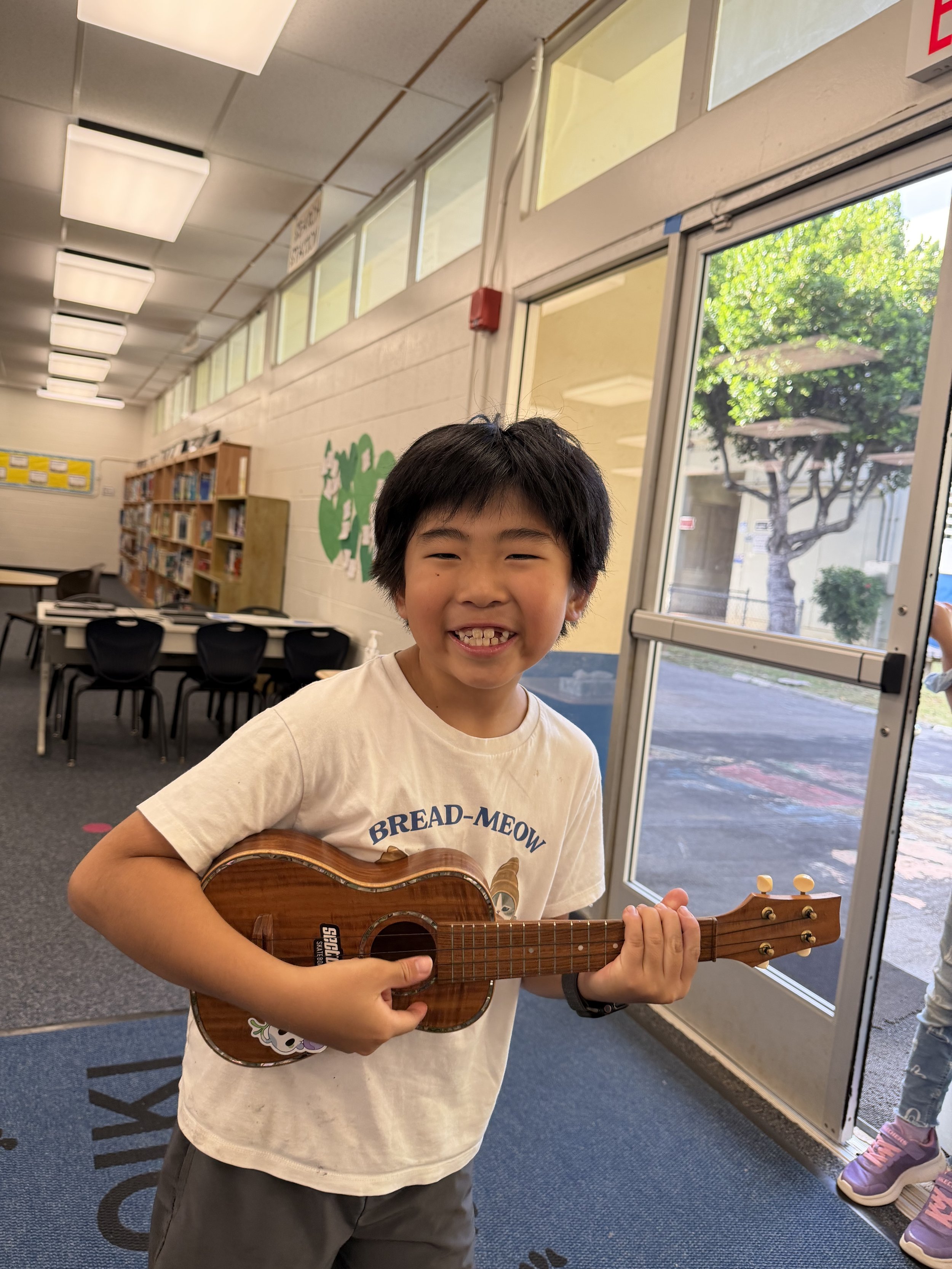 A smiling young boy playing a small wooden guitar indoors near a glass door, with bookshelves and chairs in the background.