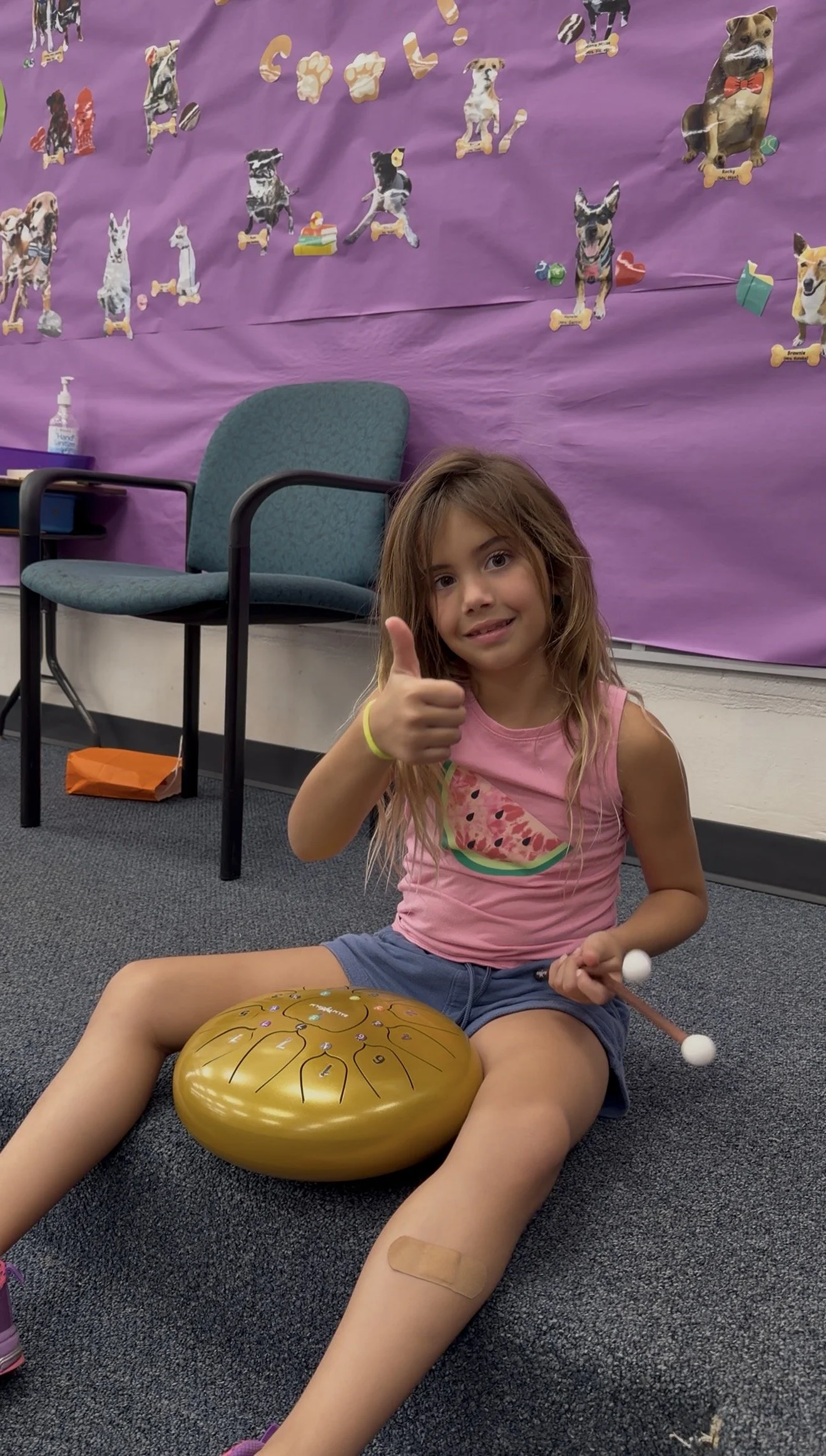 A young girl sitting on the floor giving a thumbs-up, holding mallets, with a gold metallic singing bowl featuring musical notes in front of her. She is in a room with purple wall decorations of dog-themed stickers, an office chair, and a chair with 