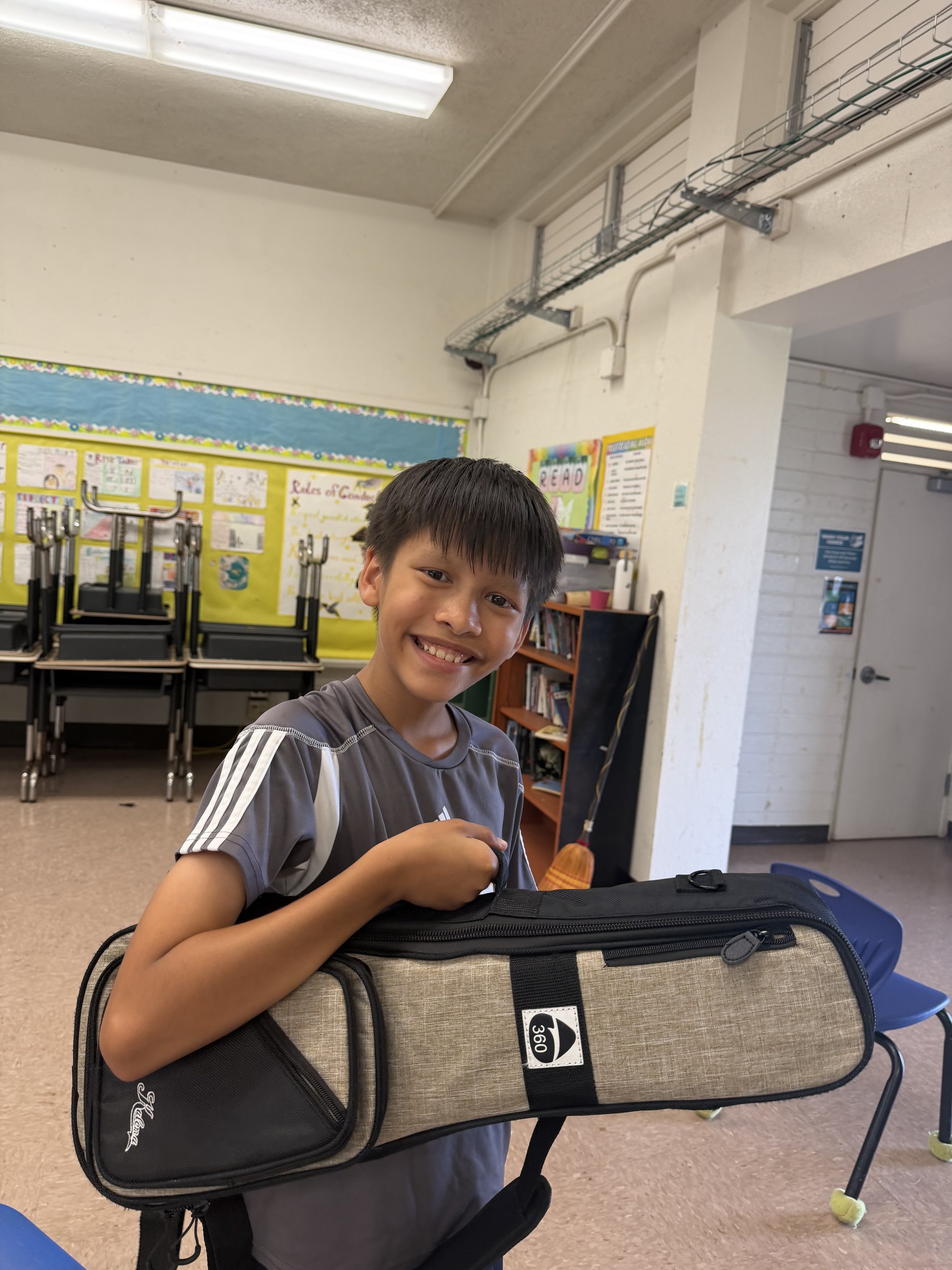 A smiling young boy with short dark hair holding a tan and black backpack in a classroom. In the background, there are folded chairs, a colorful bulletin board, bookshelves, and classroom posters.