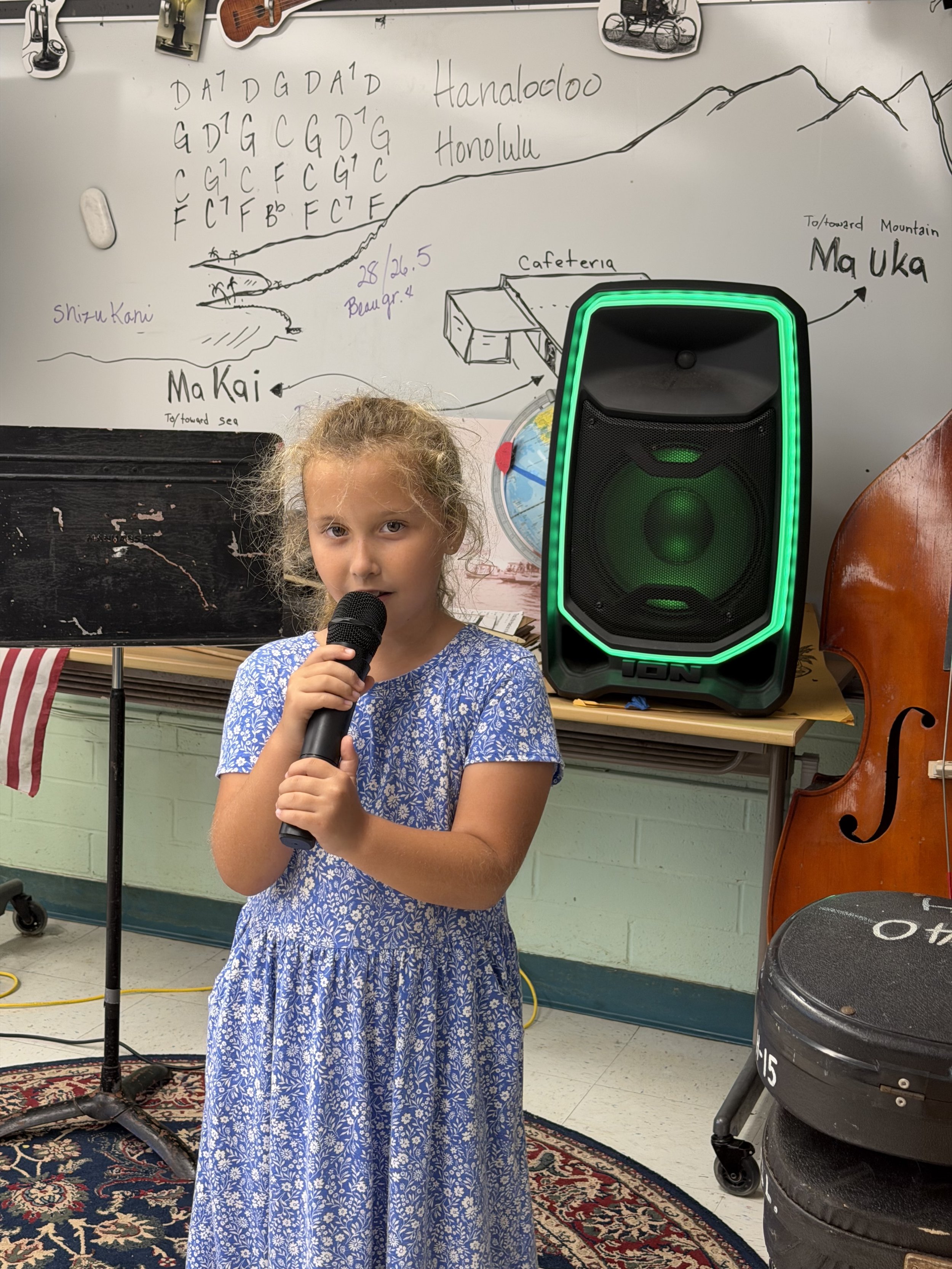A young girl with curly hair and a blue floral dress is holding a microphone in a classroom. Behind her is a whiteboard with musical notes and texts, a speaker, a globe, a music stand, and a double bass. The classroom has a patterned rug on the floor