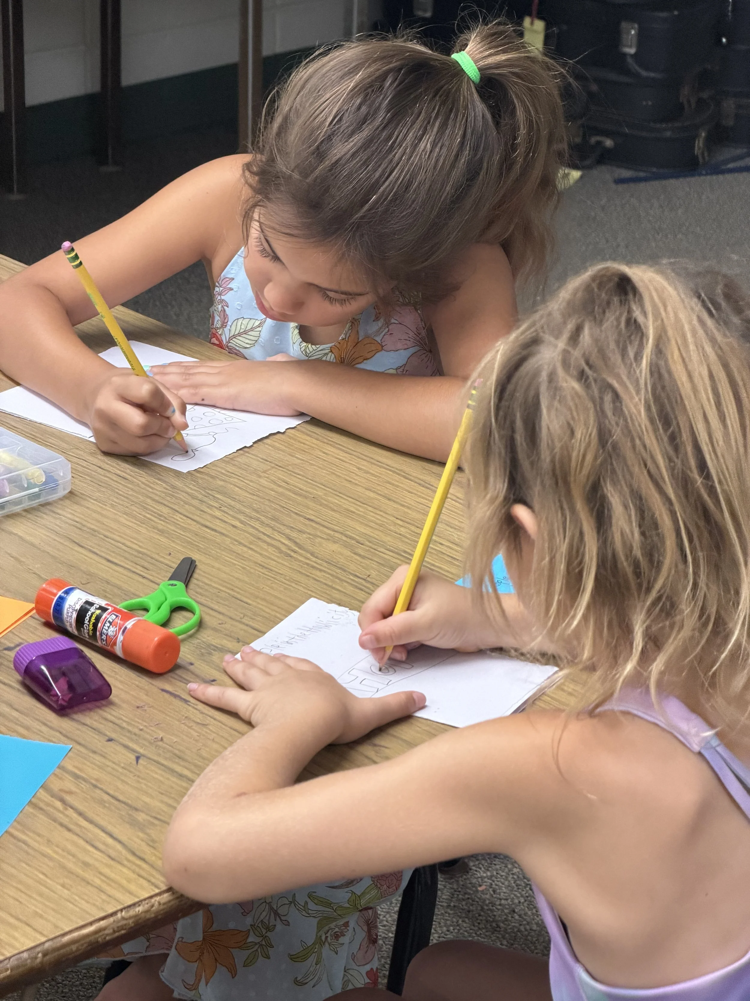 Children sitting at a table drawing and writing on paper with colored pencils, glue, and scissors nearby.