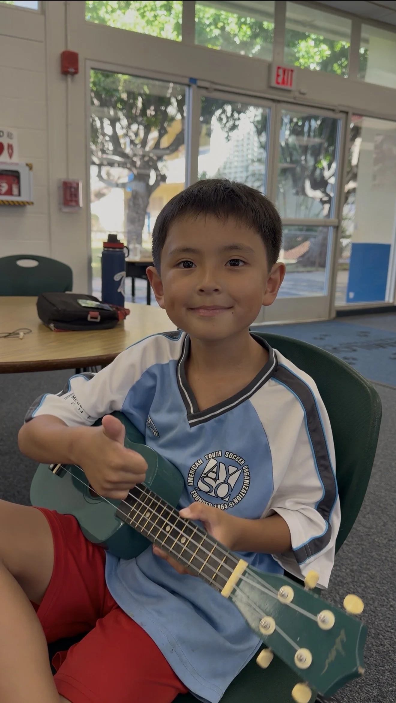 A young boy sitting at a table, holding a small blue guitar, giving a thumbs-up and smiling inside a room with large windows and green trees outside.